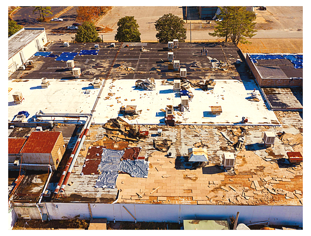 Aerial view of a rooftop with damaged sections, scattered debris, and HVAC units, with trees and a parking lot visible in the background.