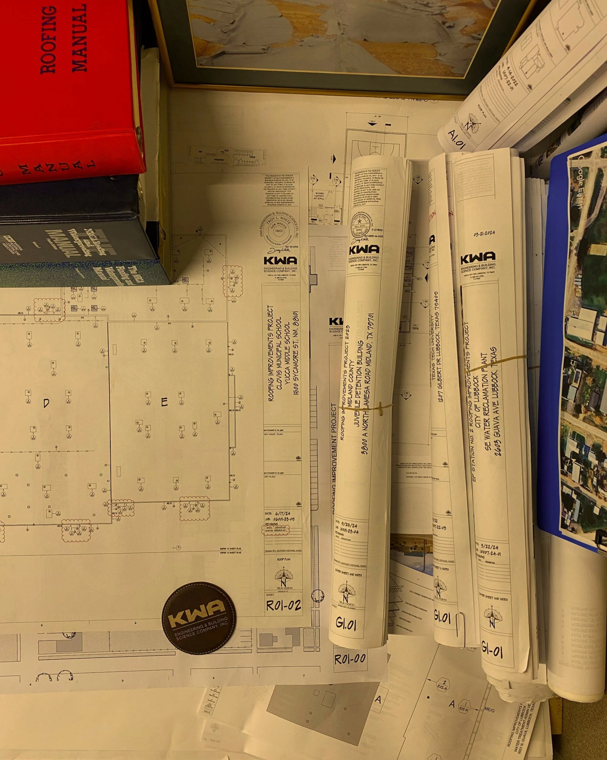 Architectural blueprints and engineering plans spread out on a desk, with a rolled-up blueprint secured with a rubber band, and a black circular sticker with 'KWA' logo.