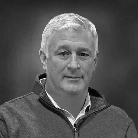 A black-and-white portrait of a middle-aged man with short, light-colored hair, wearing a zip-up jacket over a collared shirt.