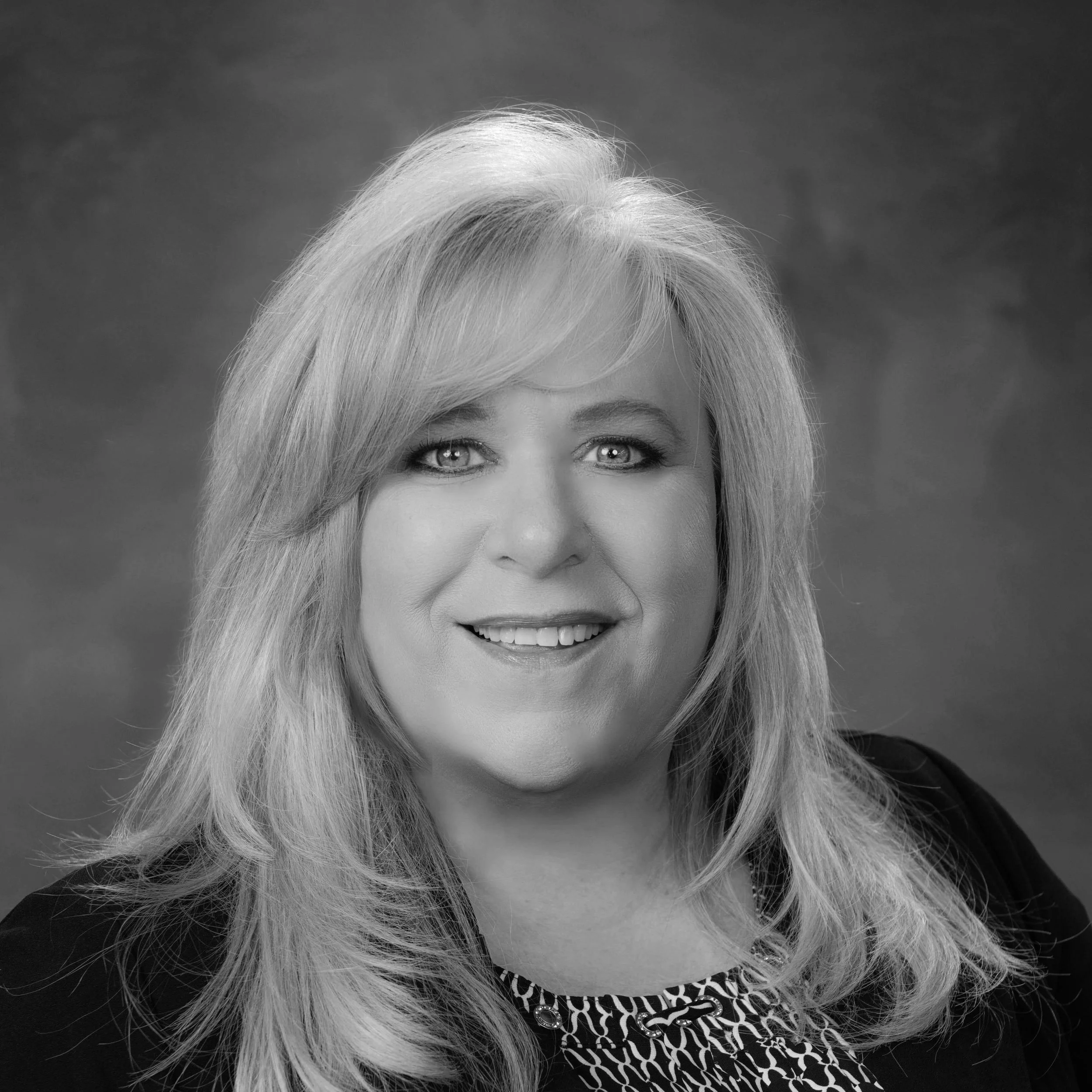 Black and white portrait of a smiling woman with long, wavy hair, wearing a patterned top and blazer, against a dark background.