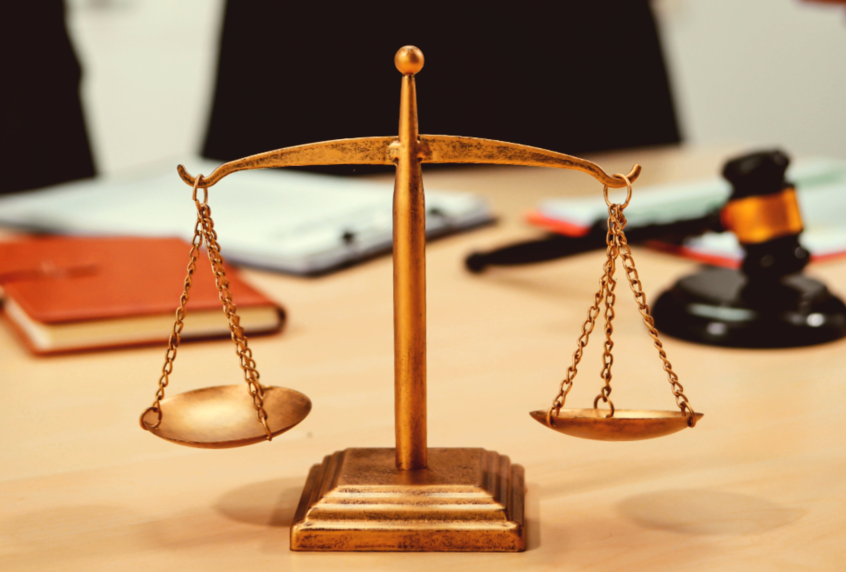 Gold-colored balance scales on a desk with legal books, papers, and a gavel in the background.