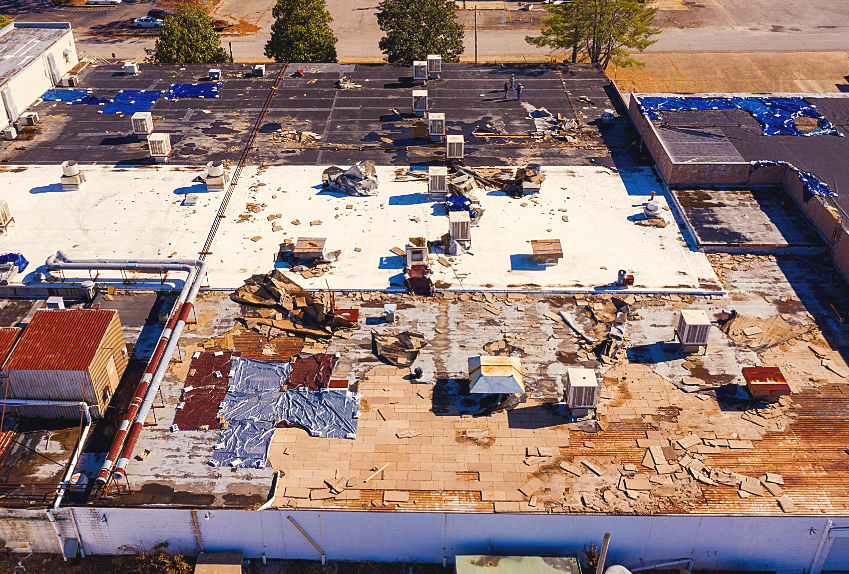 Aerial view of a commercial building roof with damage and debris, including broken tiles, torn tarps, and scattered objects, suggesting possible roof repairs or damage.