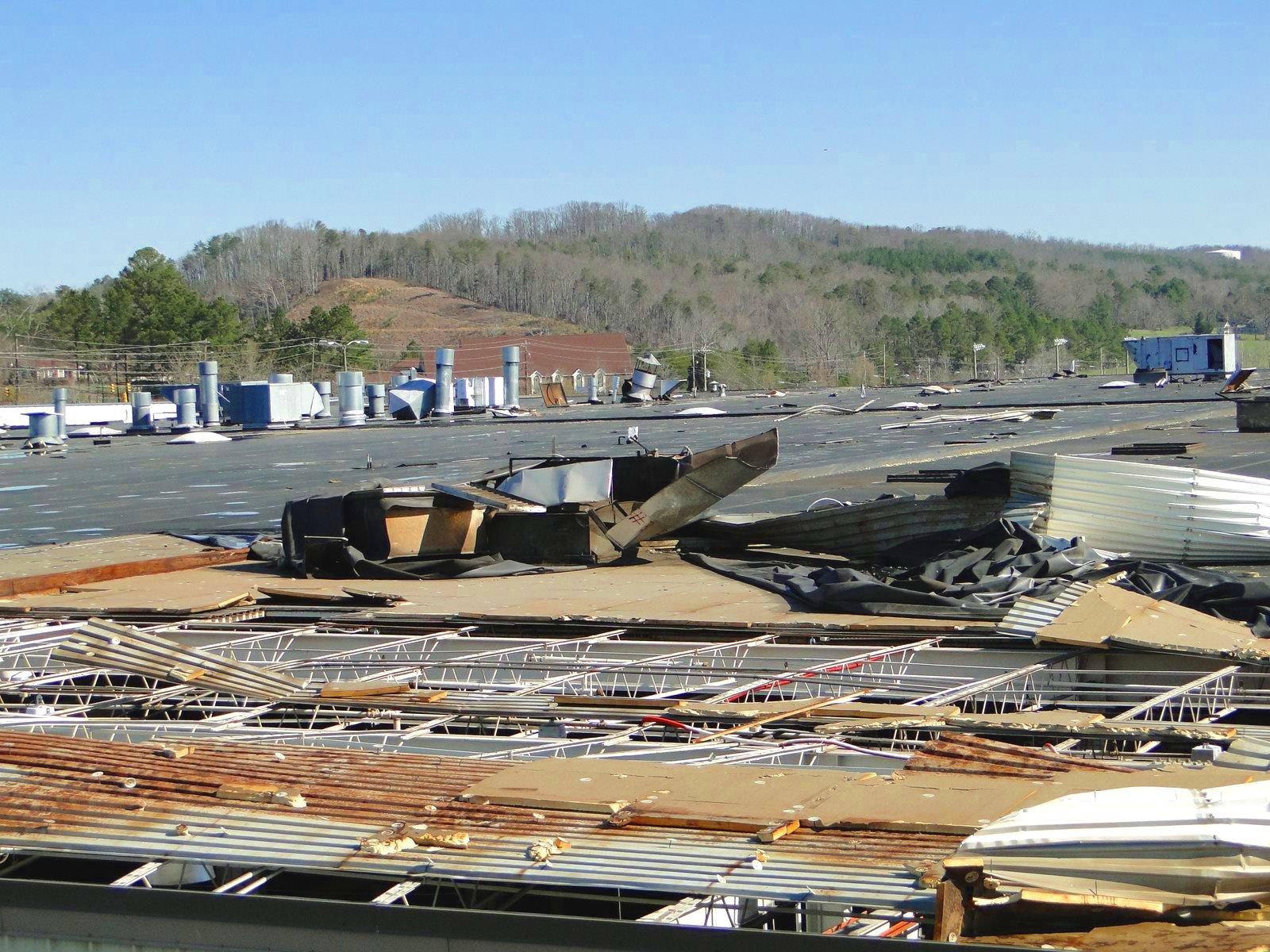 Debris and damage on a rooftop after a storm or tornado, with fallen metal roofing, shattered vents, and damaged structures under a clear sky.