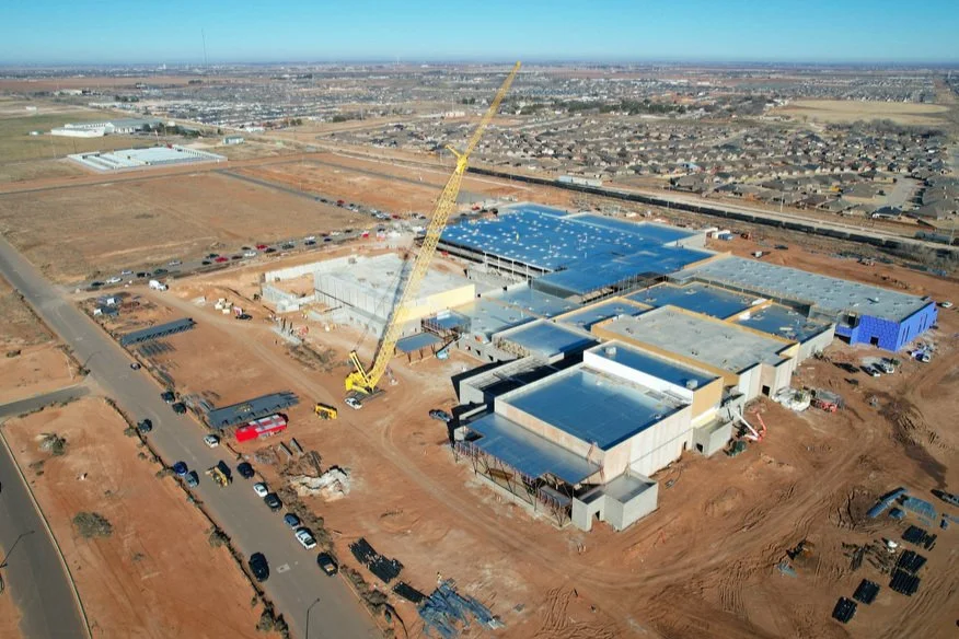 An aerial view of a construction site where a large building is under construction, with a yellow crane and construction vehicles, surrounded by dirt and parked cars.
