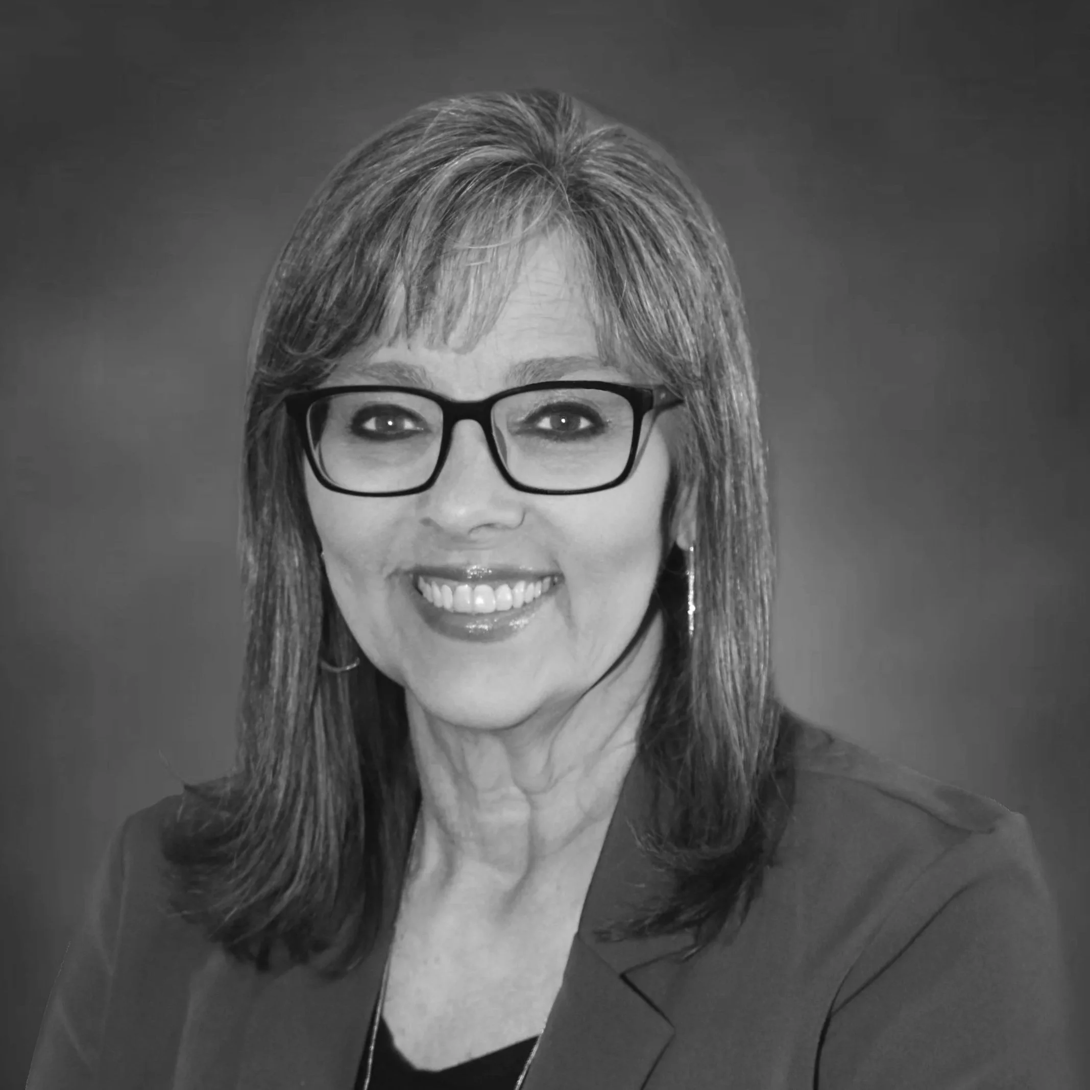 Black and white portrait of a smiling woman with shoulder-length hair wearing glasses and earrings.