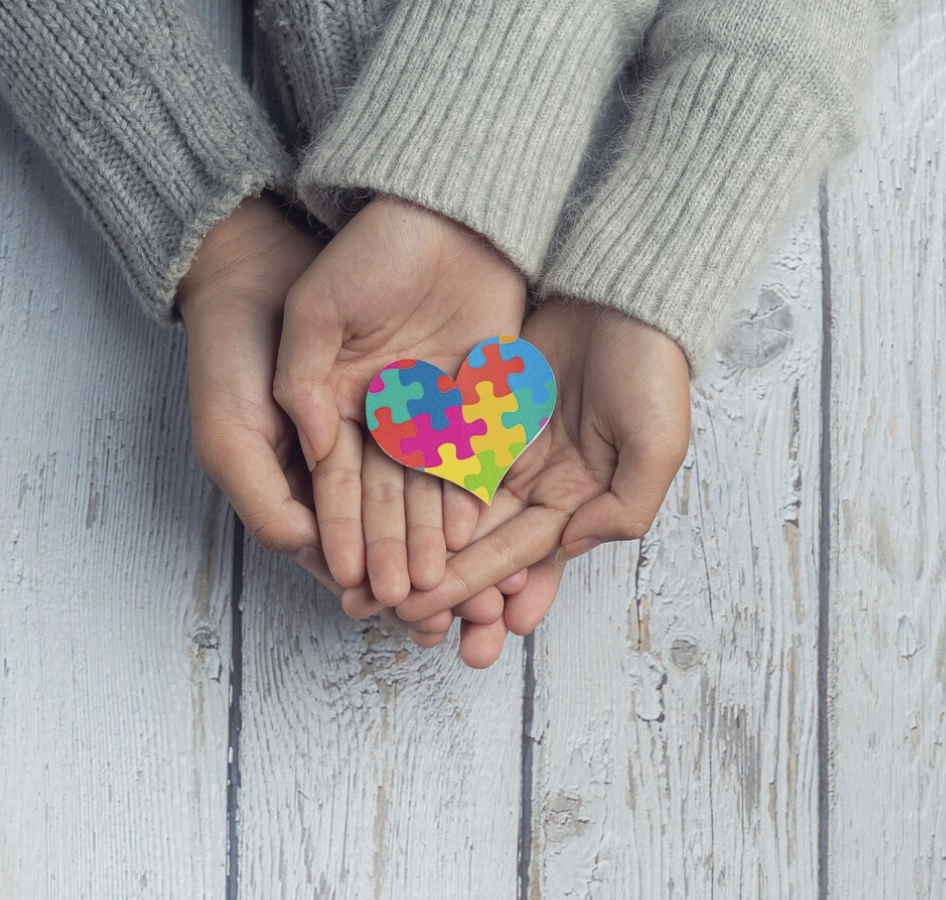 Deux mains tenant un cœur en puzzle coloré représentant l'autisme, posé sur une surface en bois blanc