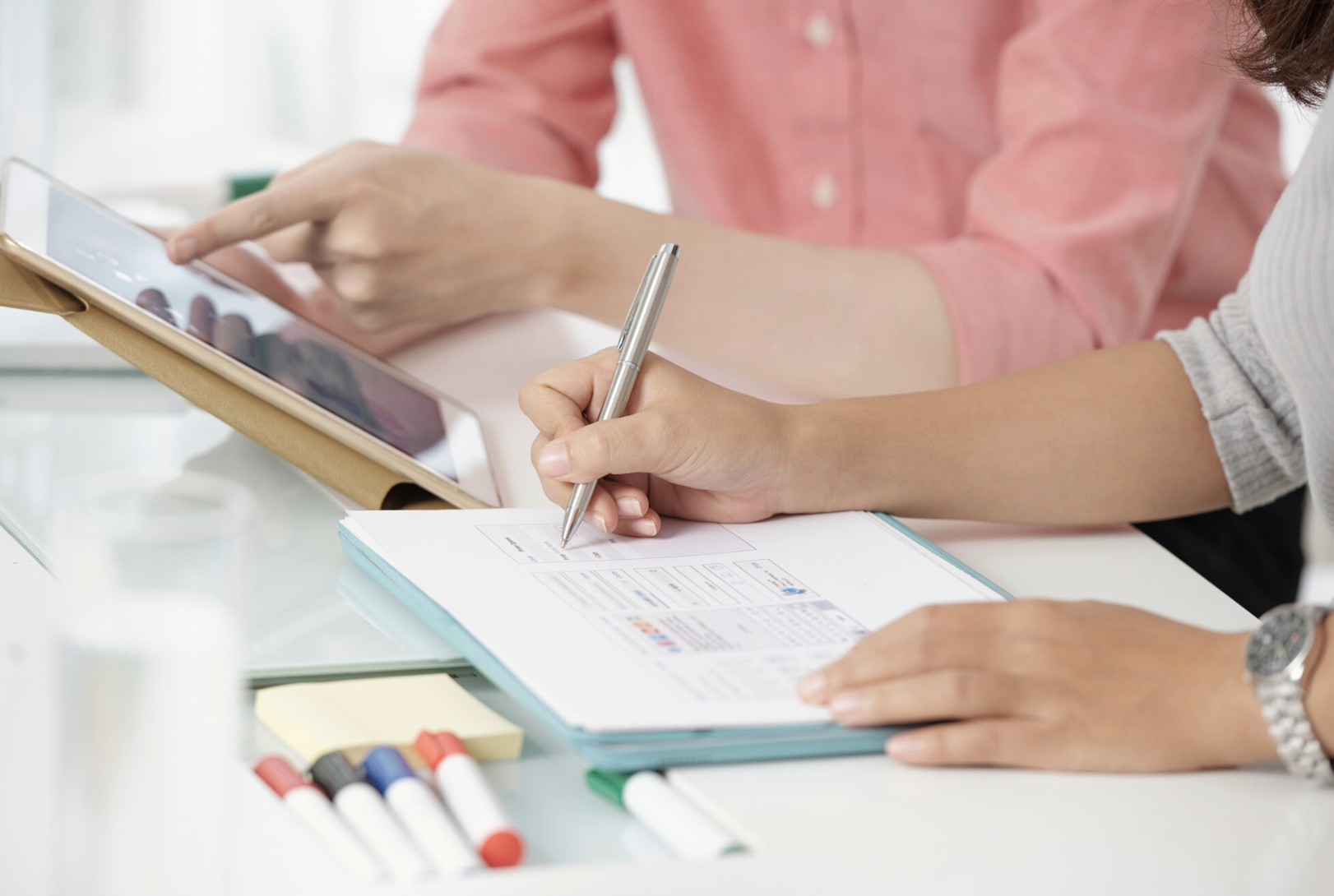 Deux personnes travaillent à un bureau, l'une écrivant sur un document avec un stylo, l'autre utilisant une tablette