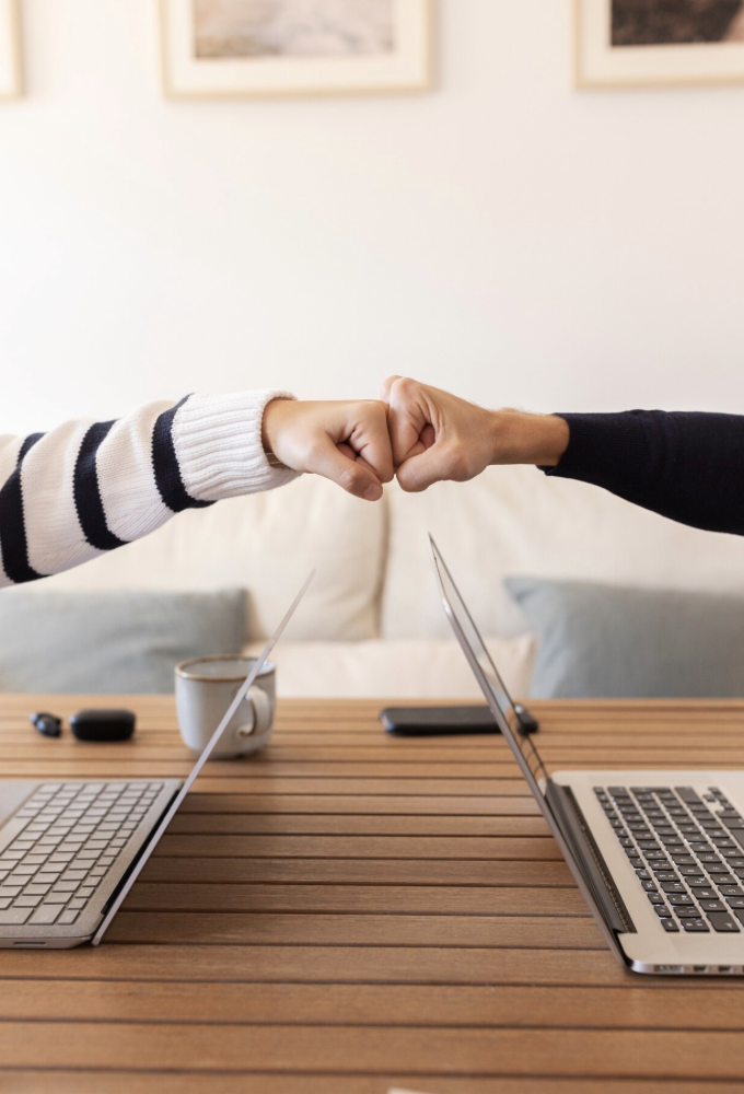 Deux personnes se donnent un high five à travers une table en bois entre deux ordinateurs portables, une tasse, une clé USB, un smartphone et une télécommande sur la table, dans un intérieur avec des tableaux au mur.