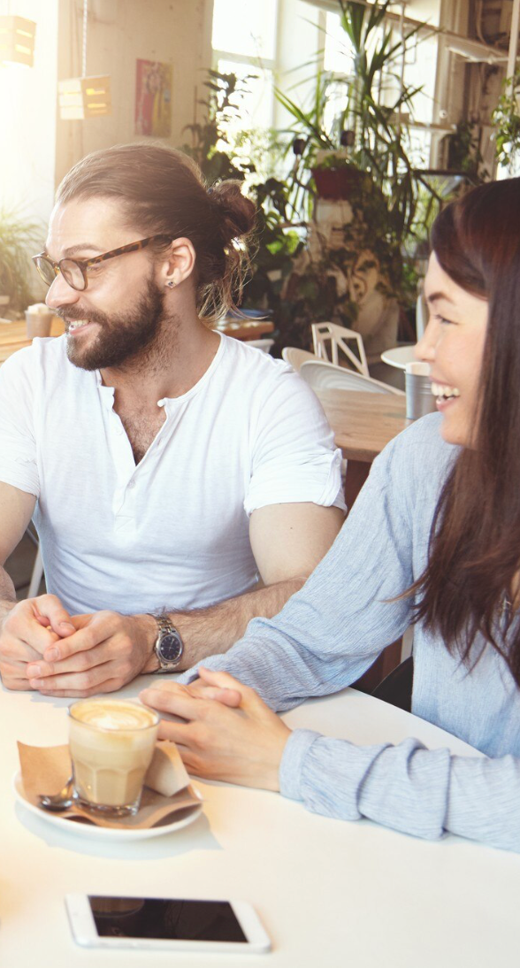 Deux personnes, un homme avec lunettes et une femme, rient et discutent dans un café lumineux avec des plantes en arrière-plan. Il y a un café latte sur la table.