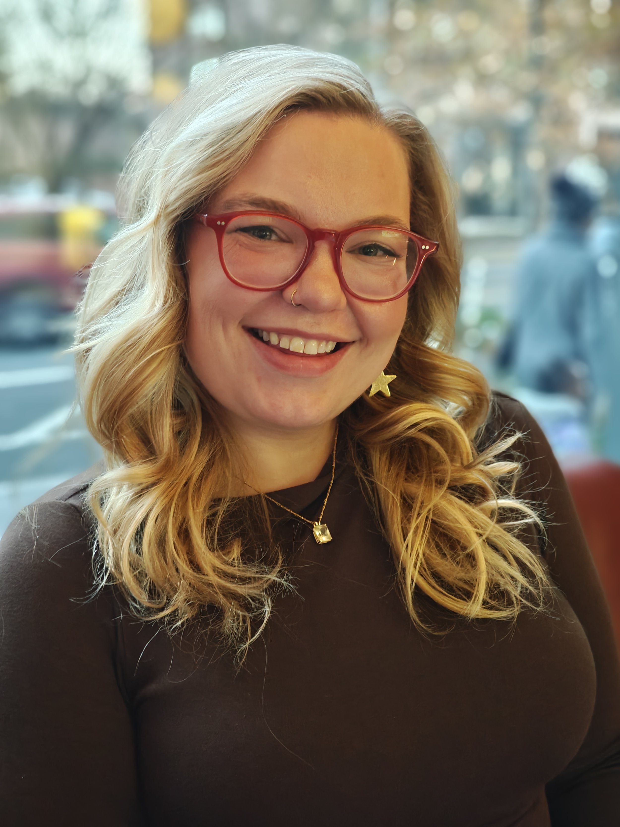 A young woman with blonde hair wearing red glasses, a dark top, star-shaped earrings, and gold jewelry. She is smiling and sitting indoors with a blurred window background showing outside scenery.