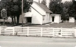 Black and white photo of a white house with a porch, a picket fence in front, and trees in the background.