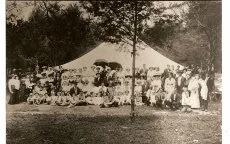 A historical black-and-white photograph of a large group of people gathered outside under a tent, with trees in the background.