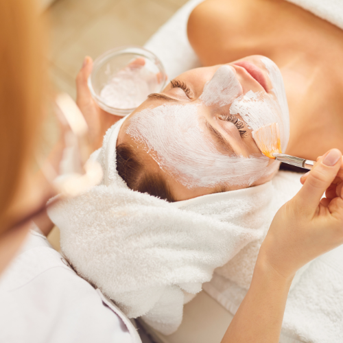 A woman lying down at a spa or skincare clinic while receiving a facial treatment, with a towel wrapped around her head and a facial mask being applied with a brush.