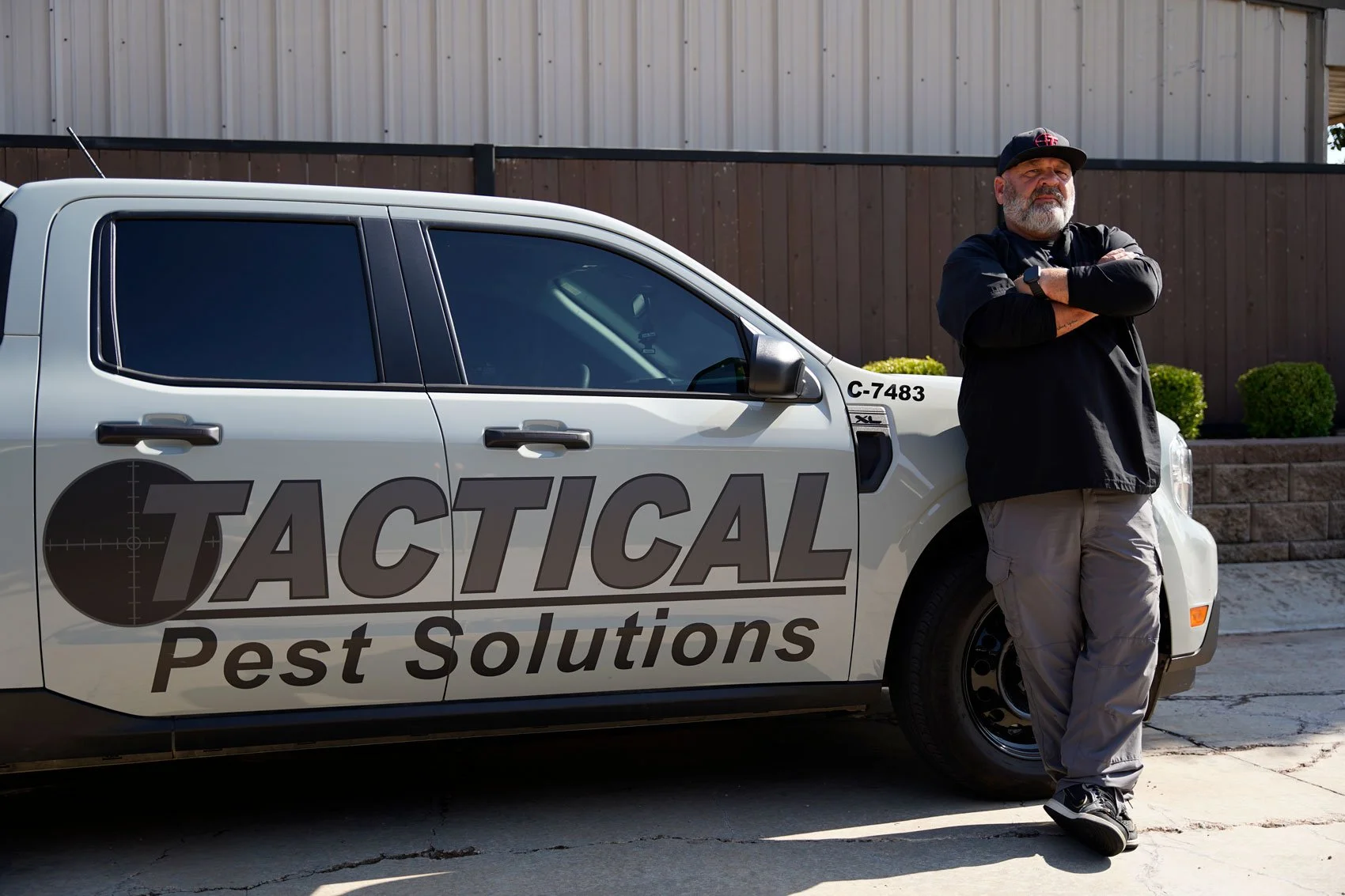 Man standing with arms crossed next to a vehicle with 'Tactical Pest Solutions' logo, outdoors in front of a wooden fence.
