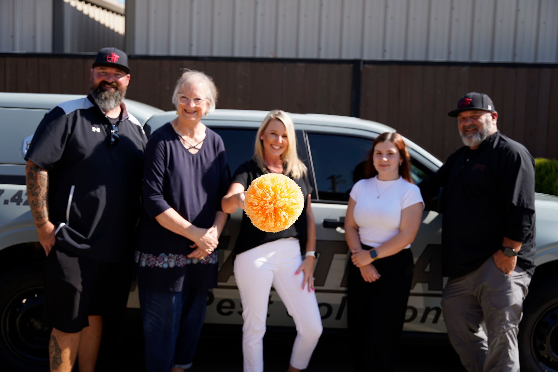 Group of five people standing in front of a police car, smiling, with a woman in the center holding an orange pom-pom decoration.