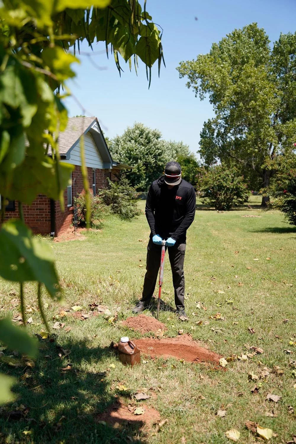 A person in black clothing and gloves digging a hole in a grassy yard with a shovel, surrounded by green trees and a house in the background on a sunny day.