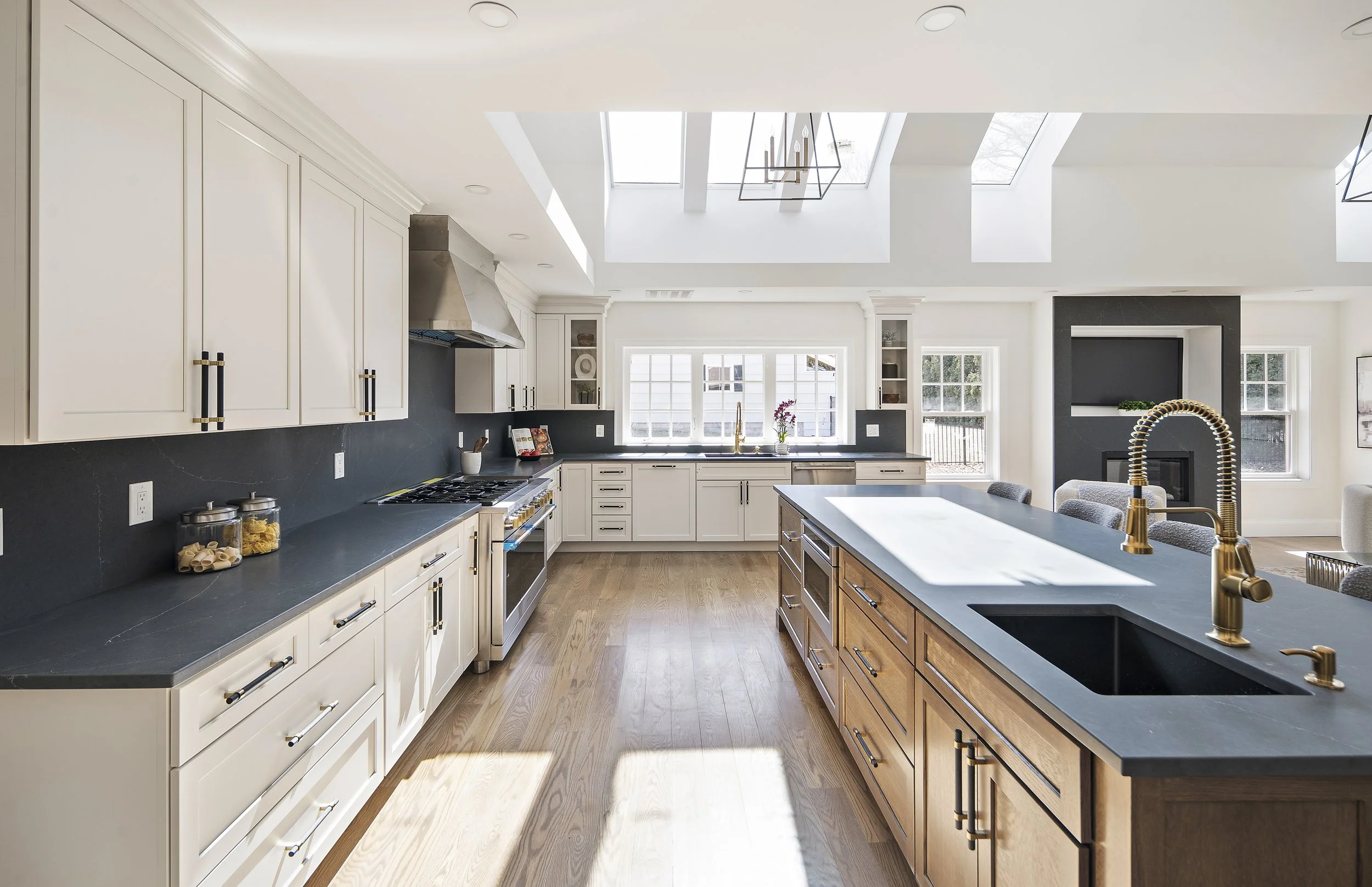 Modern kitchen with white and wood cabinets, large window, skylights, black backsplash, stainless steel appliances, and a central island with a gray countertop and gold faucet. One of our projects in Andover, MA.