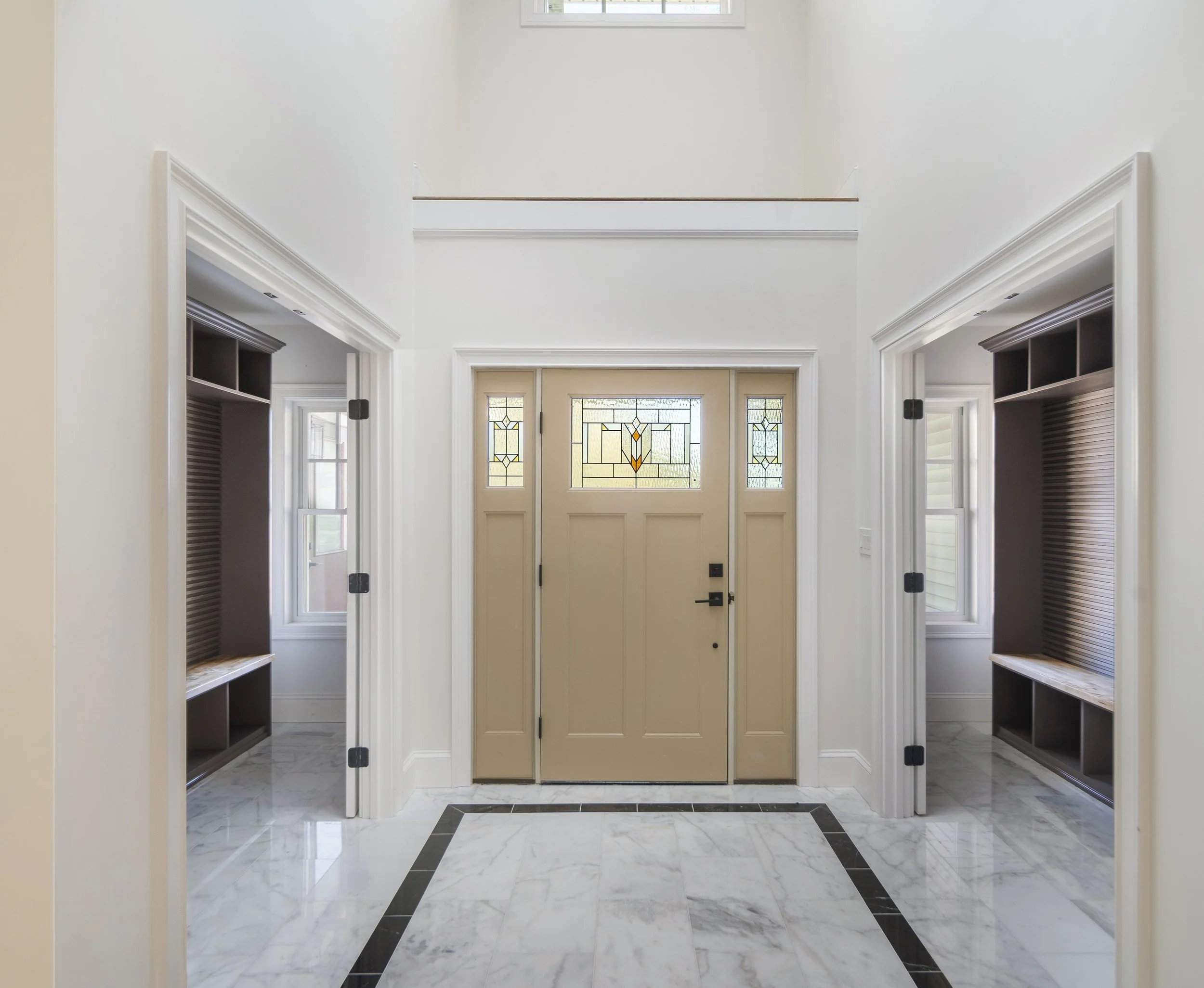 Entryway with a central beige door featuring stained glass panels, flanked by two side windows, and two open built-in cubby storage areas with dark wood finish, situated on marble flooring with a black border.