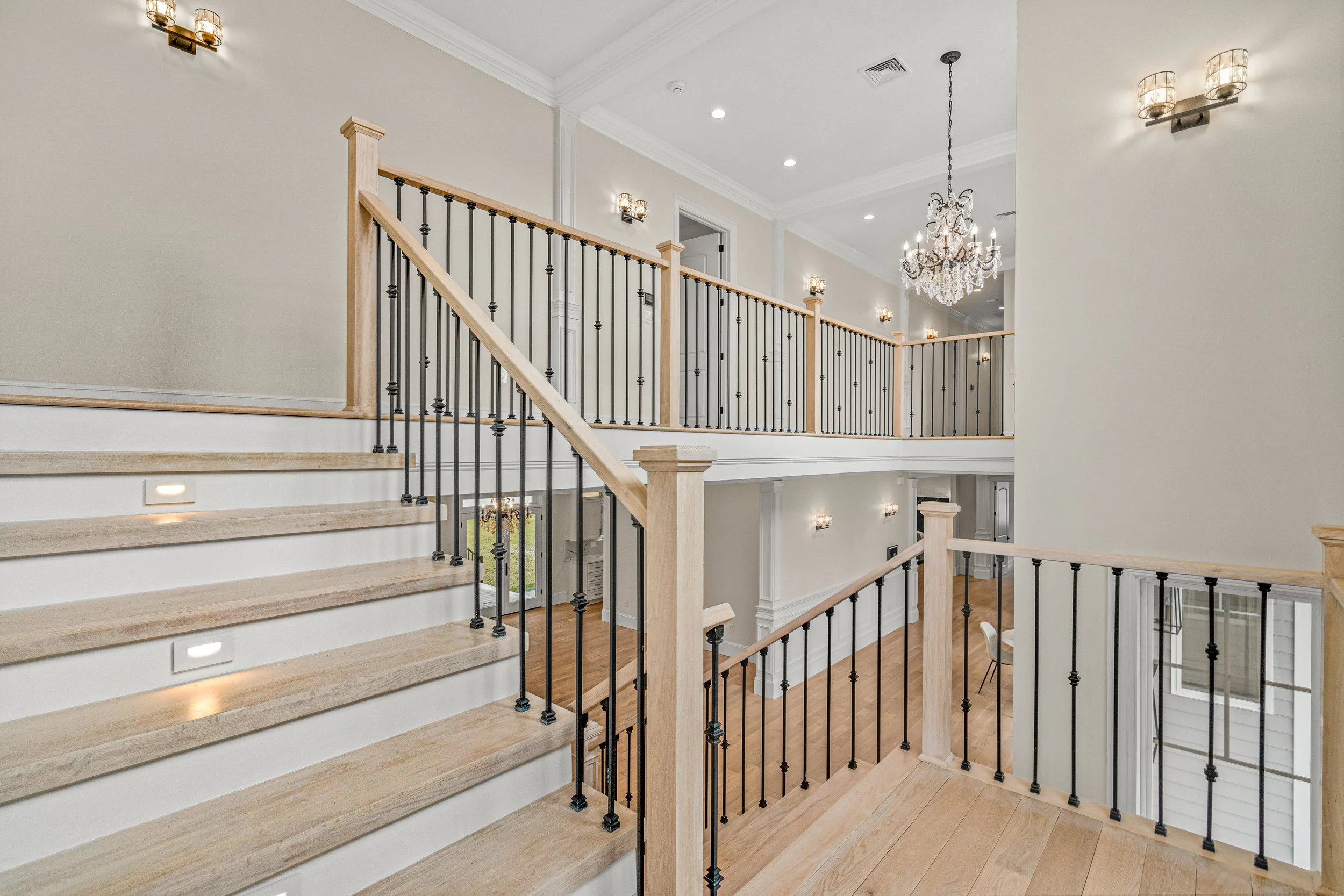 View of a spacious staircase and upper hallway in a modern home with white walls, wooden flooring, decorative chandeliers, and wall-mounted lights. One of our projects in Lexington, MA.
