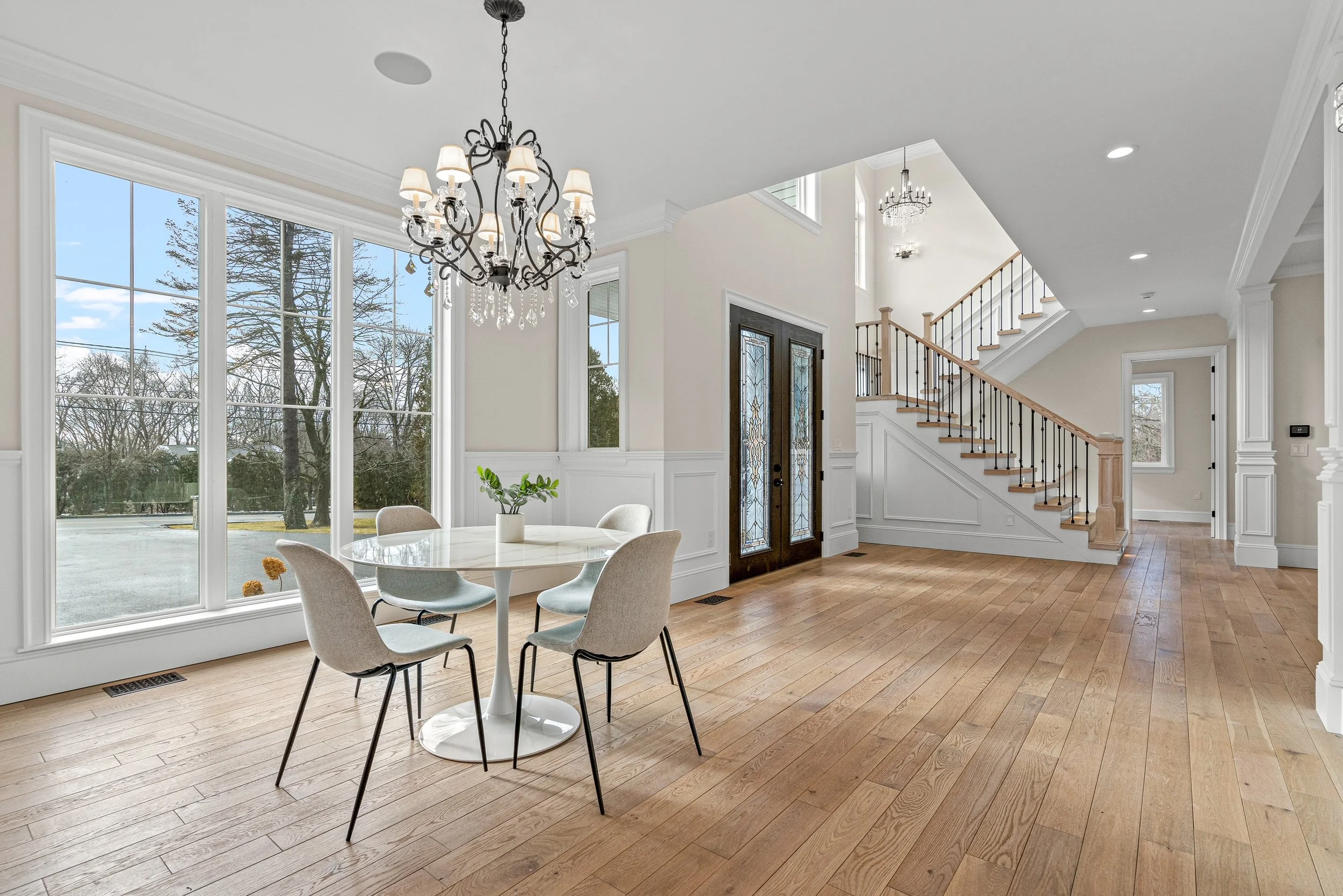 Bright living room with large windows, a round dining table with six chairs, a chandelier, stairway, and hardwood floors.