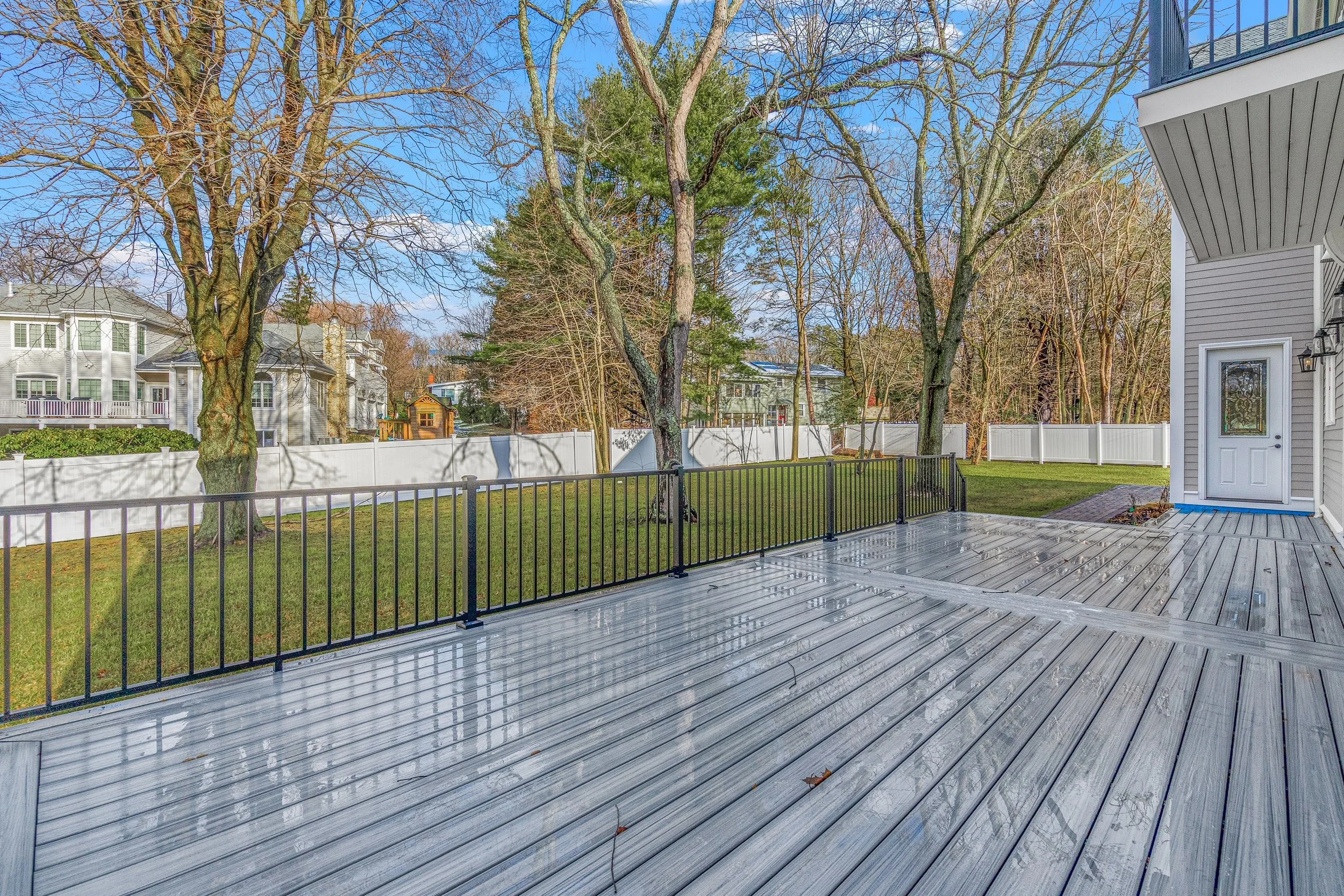 Backyard with a wooden deck, green grass, trees, and a white fence surrounding the yard.