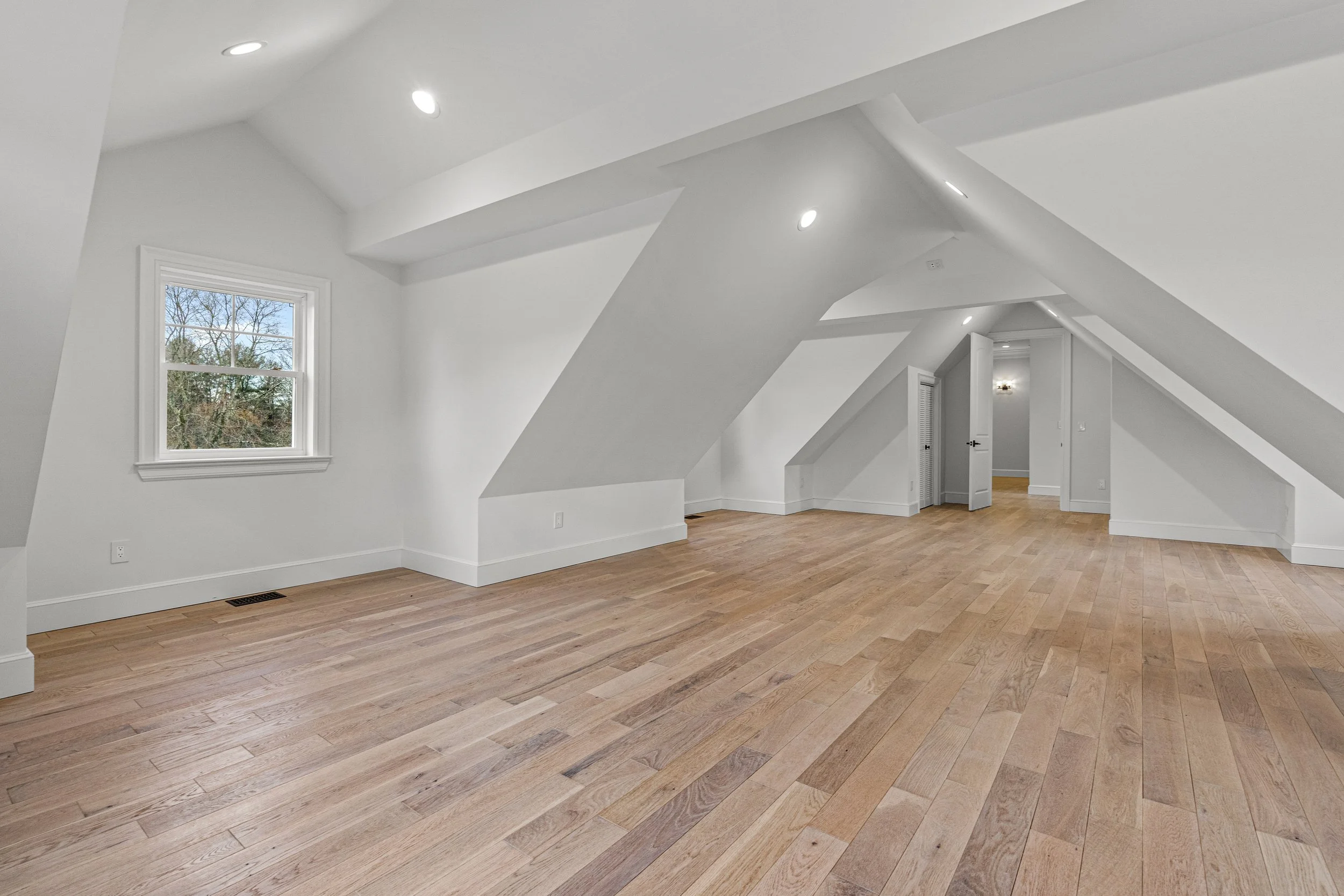 Empty room with white walls, sloped ceilings, hardwood floors, a window showing trees outside, and built-in window and door trim.