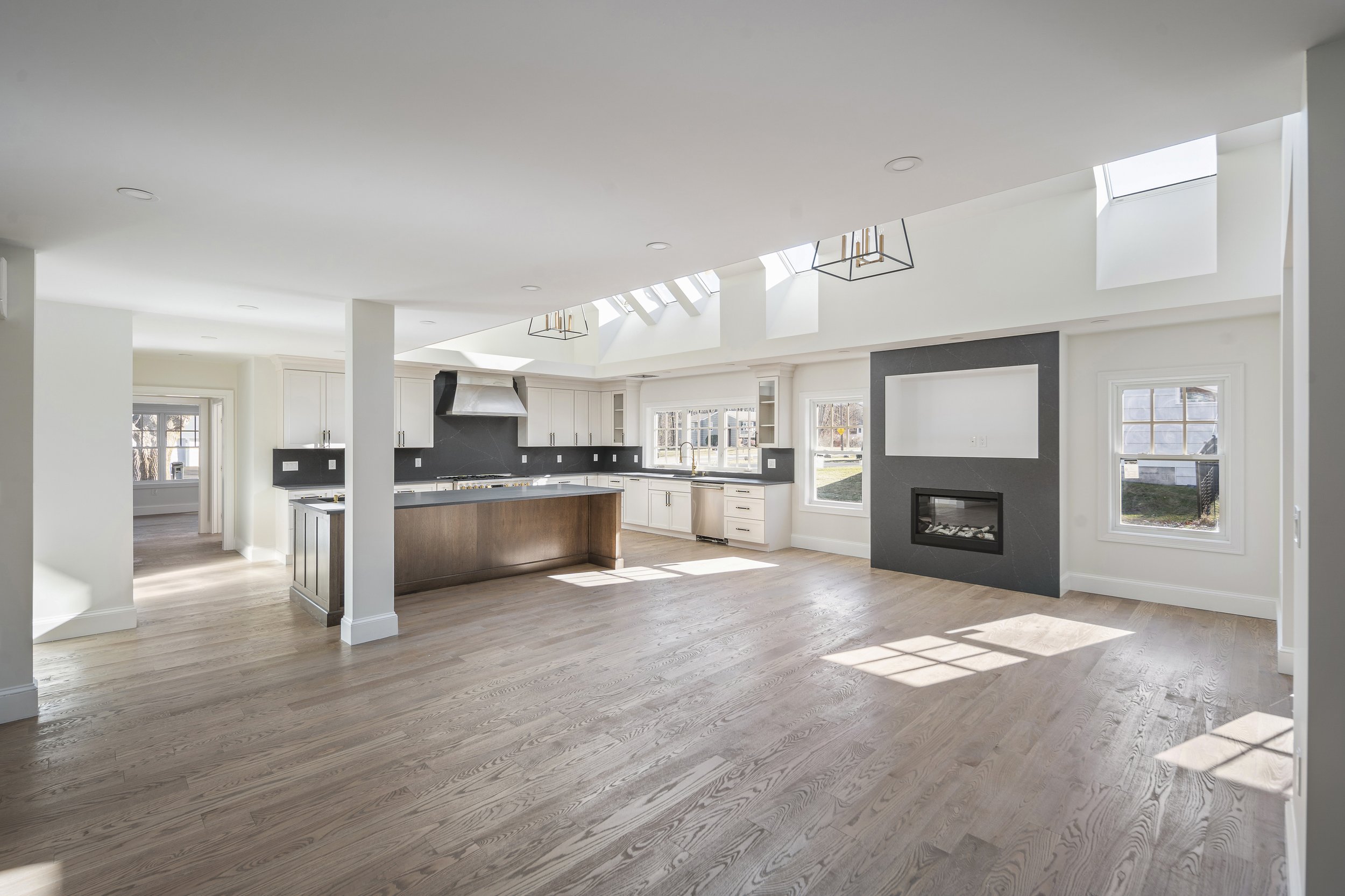 Open-concept living space with hardwood floors, white walls, large windows, a black fireplace, and a kitchen with white cabinets, black countertops, and an island with wooden paneling.