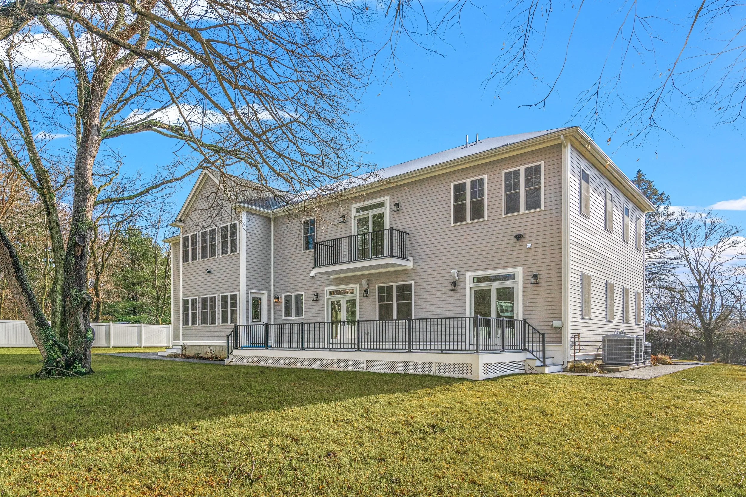 A large two-story house with beige siding and white trim, multiple windows, and a large wooden deck with black railing in the backyard. Bare trees and a white fence are visible in the background, and the sky is partly cloudy with some blue showing.
