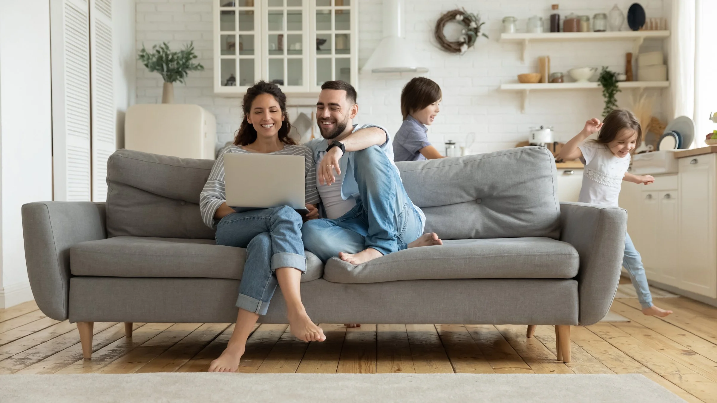 A family of four spending time together in their living room during daytime. A woman and a man sit closely on a gray sofa, smiling at a laptop. Two children, a boy and a girl, are in the background near the kitchen, playing and laughing.
