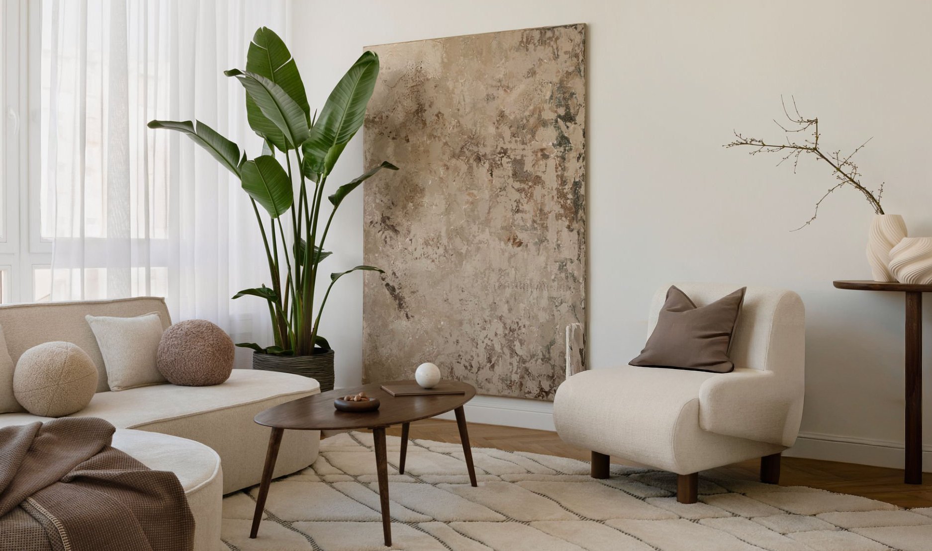 Living room with a beige sofa adorned with round cushions, a wooden coffee table, a white armchair with a brown pillow, a large green potted plant, a textured wall art, and decorative branches on a side table, with natural light coming through sheer curtains.