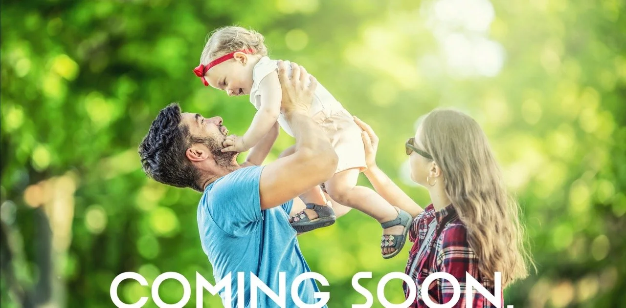 A family of three enjoying outdoors in a green park, with the father lifting the young daughter into the air and the mother smiling at them.