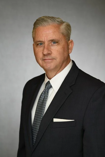 A professional headshot of a middle-aged man with blonde hair, wearing a dark suit, white shirt, and patterned tie, standing against a gray background.