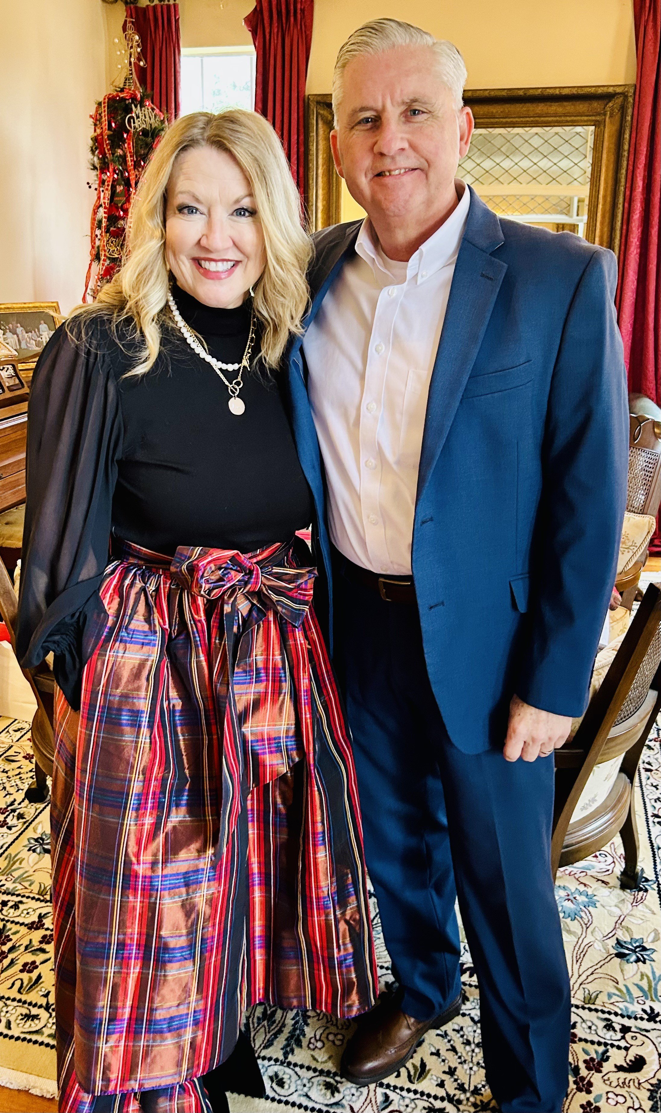 A smiling woman with blonde hair in a black top and plaid skirt stands next to a man in a blue suit in a warmly decorated room with Christmas decorations.