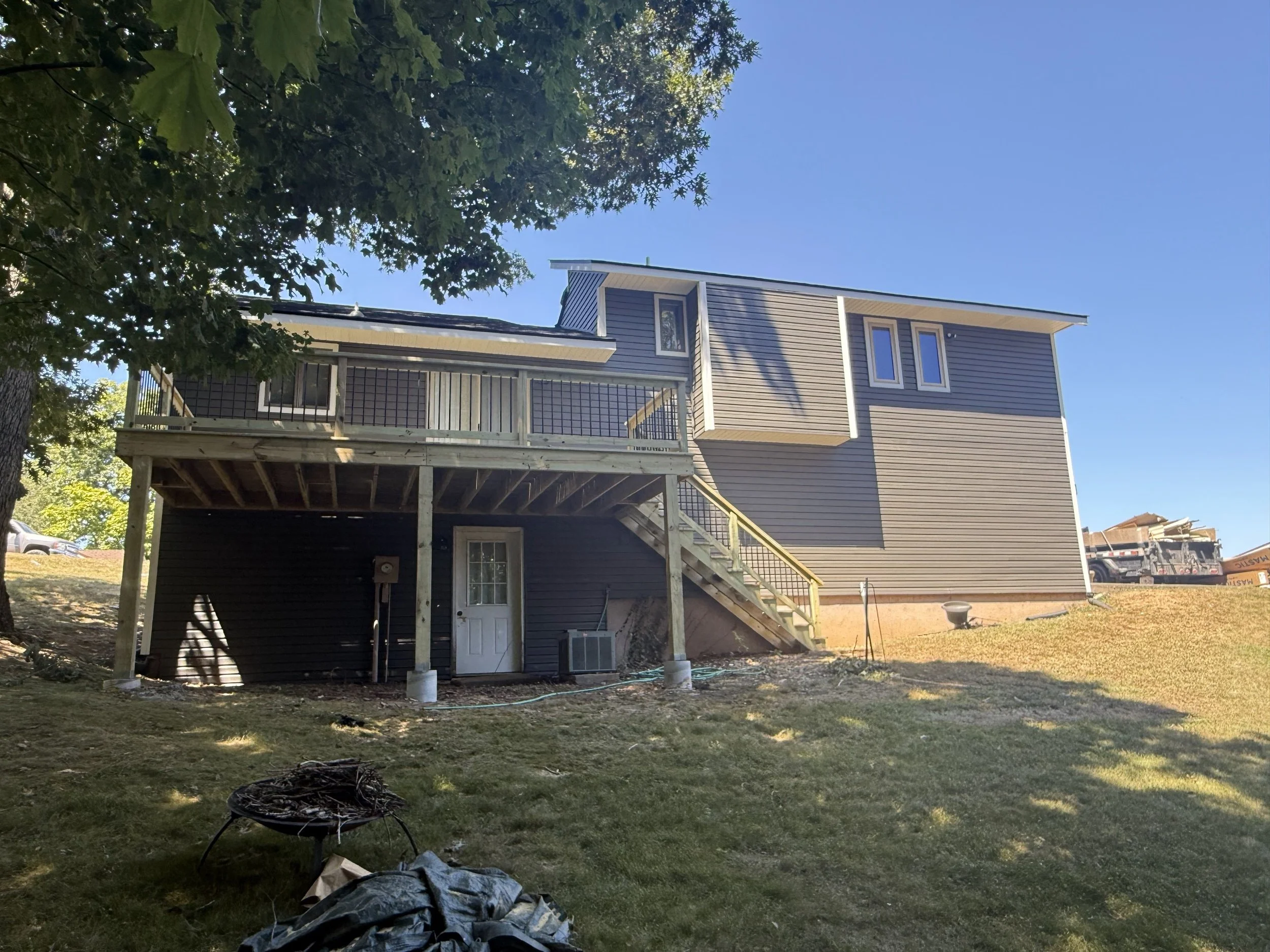 A two-story house with gray siding, a wooden deck on the upper level, and stairs leading down to the yard. The yard has grass, trees, and some construction equipment visible in the background.