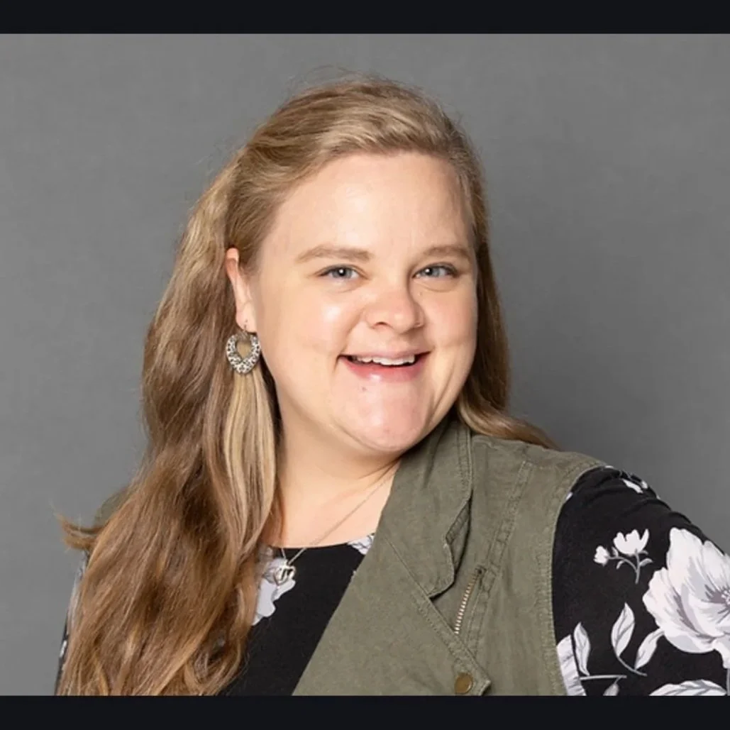 A man sitting on a beige bench against a cream-colored wall, wearing a blue short-sleeve sweater and beige pants, looking to his left with shadow cast on the wall behind him.