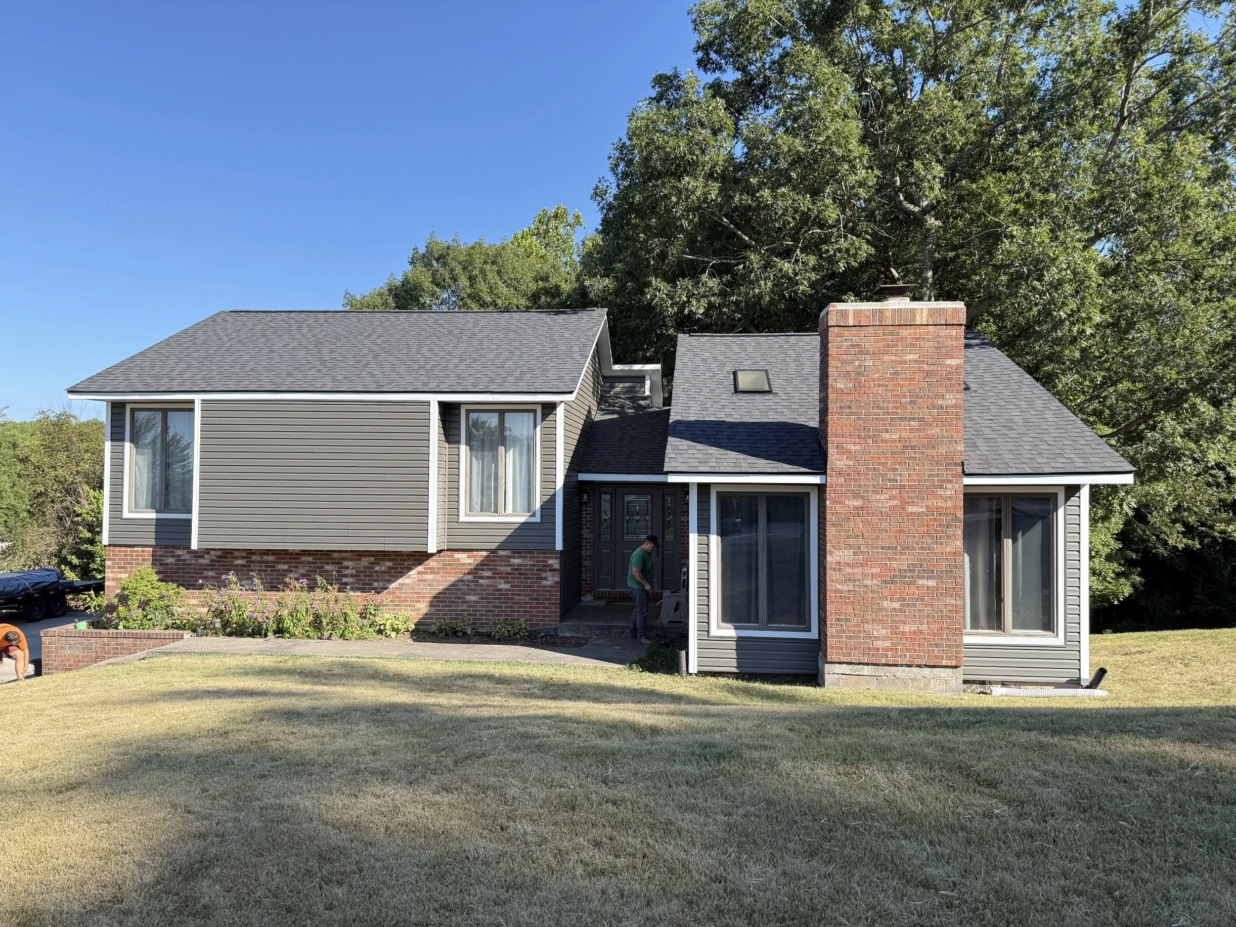 A two-story house with gray siding, brick accents, and a large brick chimney. The house has large windows and is surrounded by a landscaped yard with green grass and bushes. A person is working near the front door, and trees are in the background under a clear blue sky.