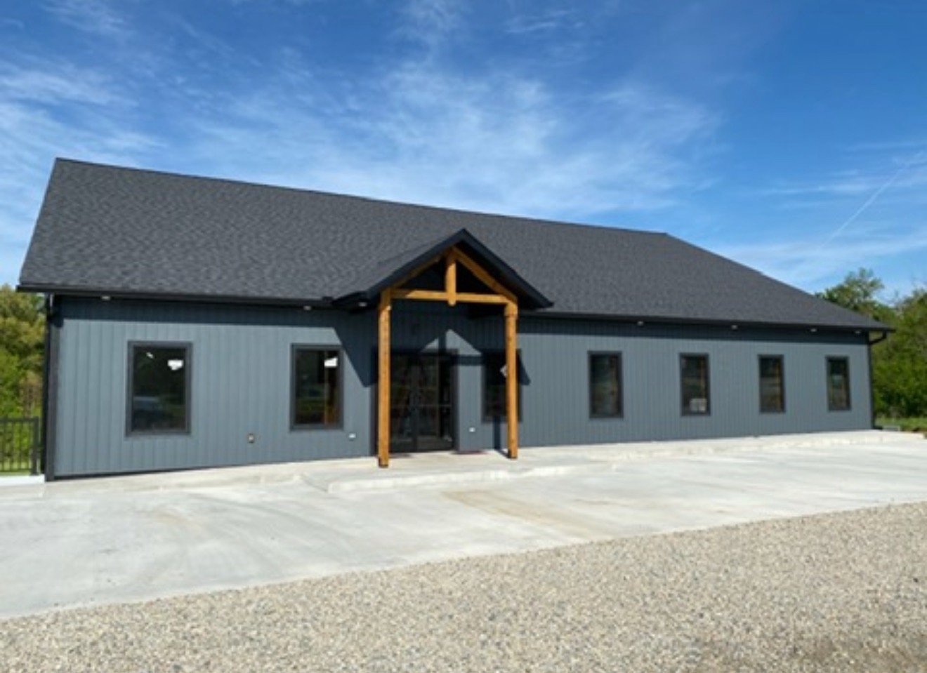 A modern, gray, single-story building with a gabled roof and a small covered entrance supported by wooden beams. The building has five windows and is surrounded by a concrete parking area with a gravel section in front. The sky is blue with some wispy clouds.