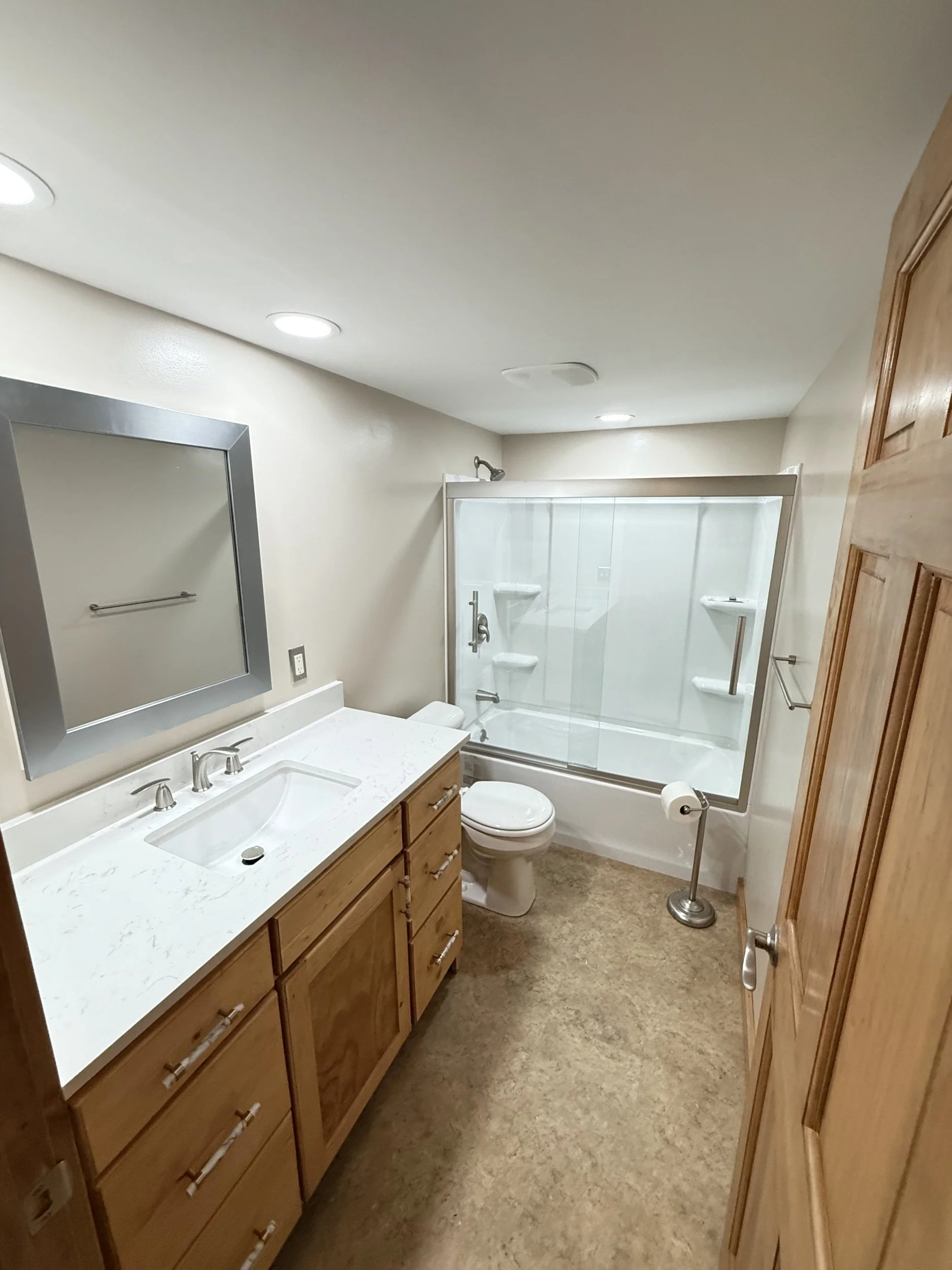 A bathroom with a wooden door, wooden vanity with a white countertop, a large mirror, a toilet, and a shower with glass doors and built-in shelves.