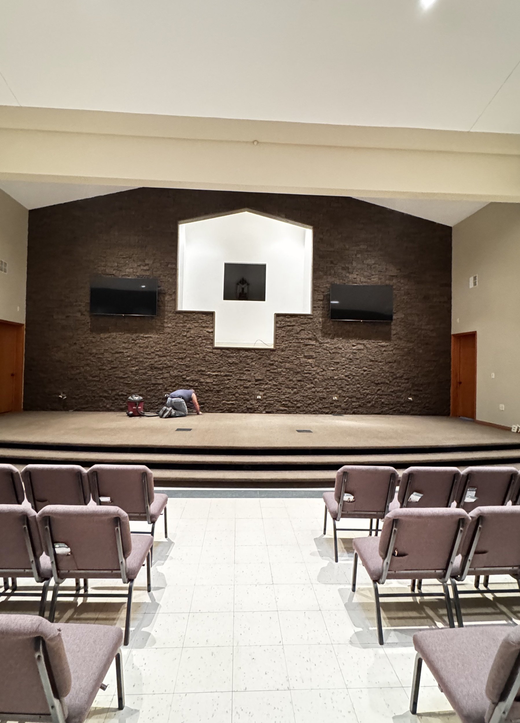 An indoor auditorium with a small stage, brown textured wall, two TV screens, and rowed chairs ready for an event.