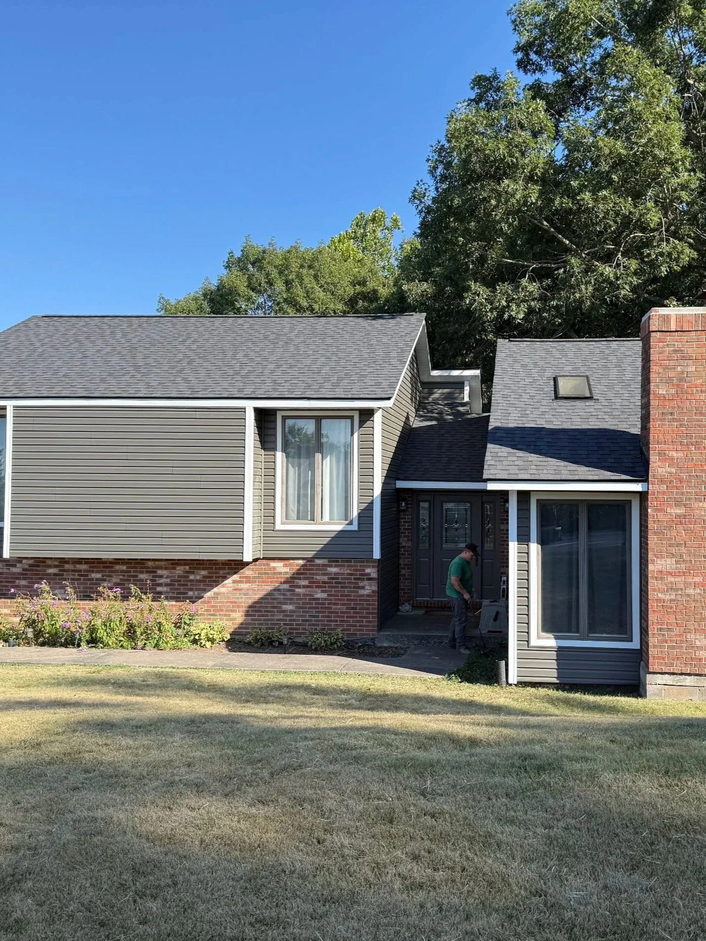 A man standing outside a two-story house with a backyard, gray siding, brick accents, and a large window, with trees and a clear blue sky in the background.