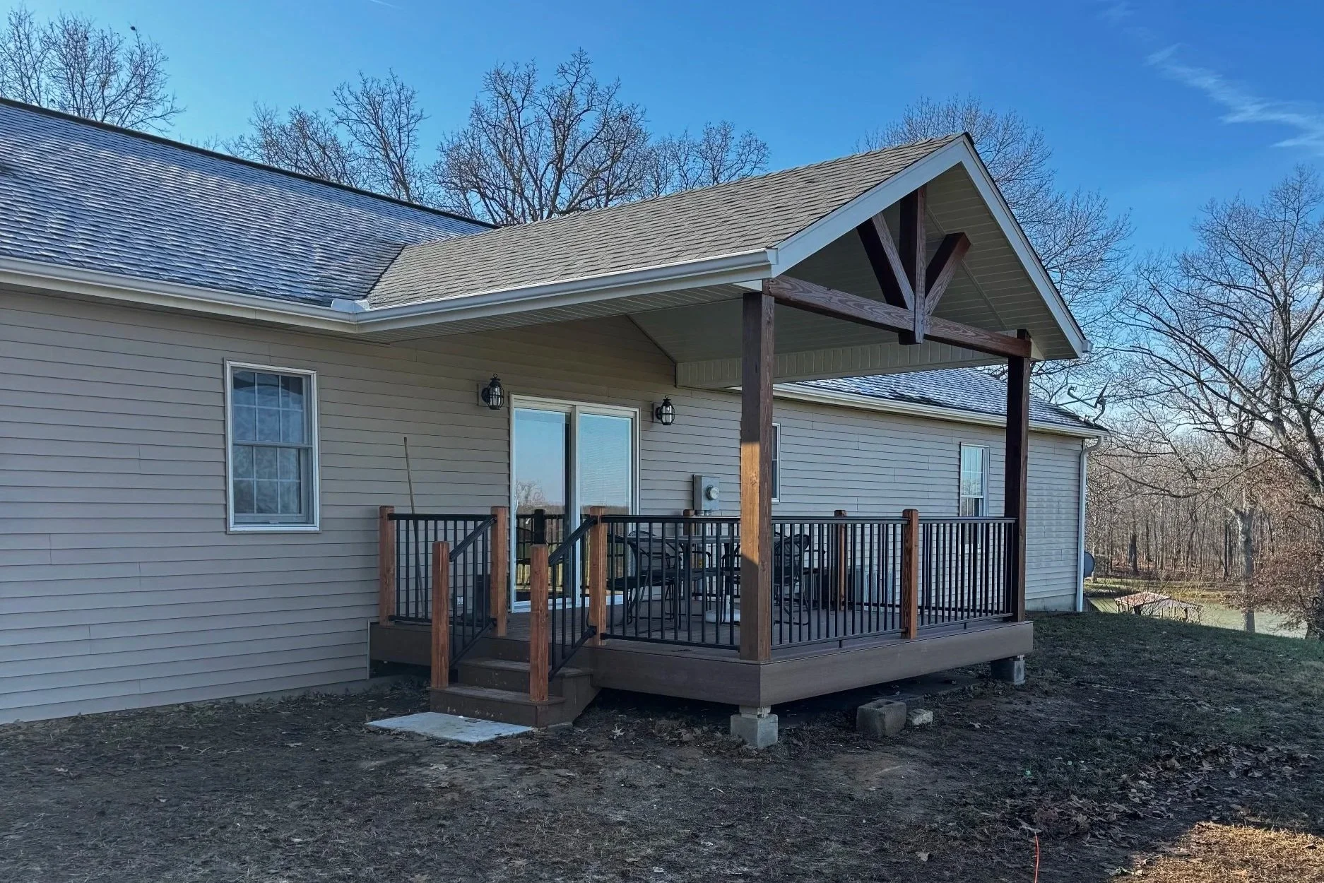 Back view of a house with a wooden deck and black railing, sliding glass door, and two wall-mounted lanterns, surrounded by leafless trees and a clear blue sky.
