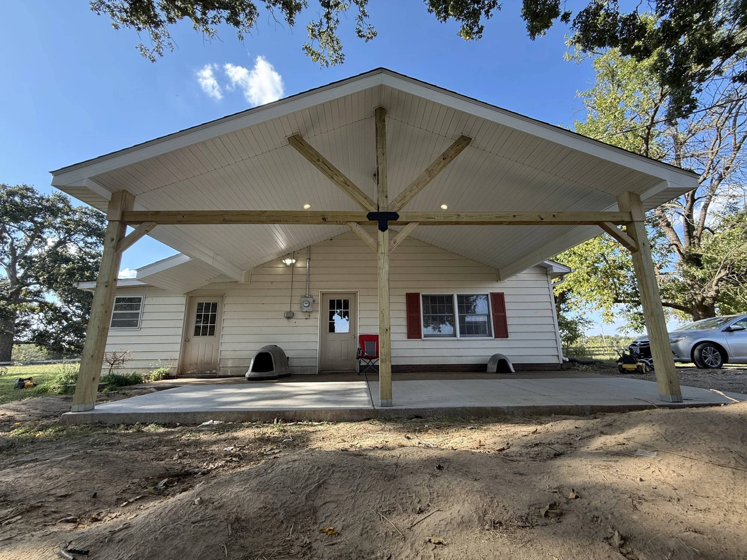 A house with a newly constructed covered patio, featuring wooden support beams, white siding, and red shutters, with a concrete floor and a car parked nearby.