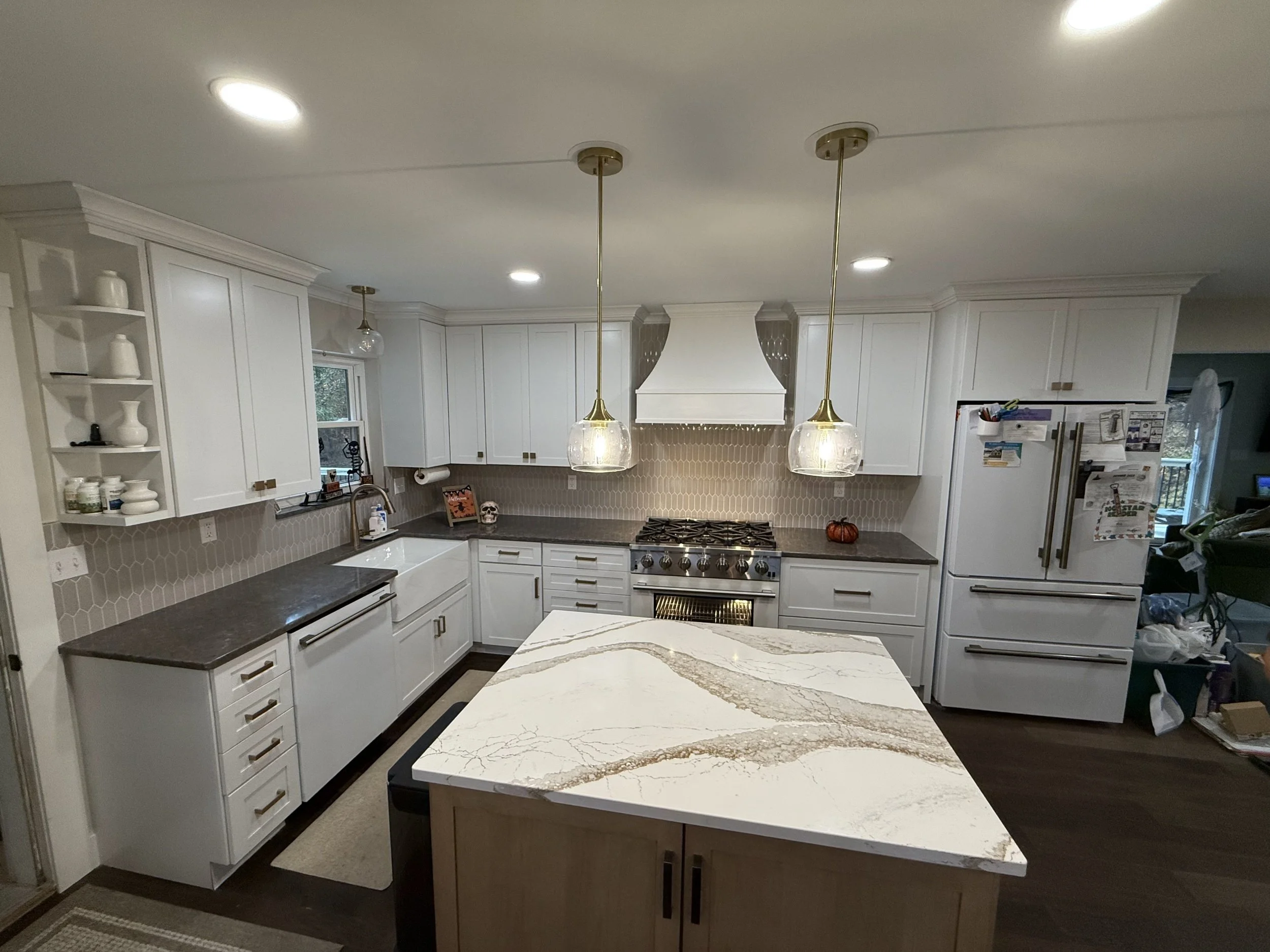 Modern white kitchen with dark counters, a island with a white and brown marble top, and gold and glass pendant lights, with a stove, refrigerator, and open shelving.