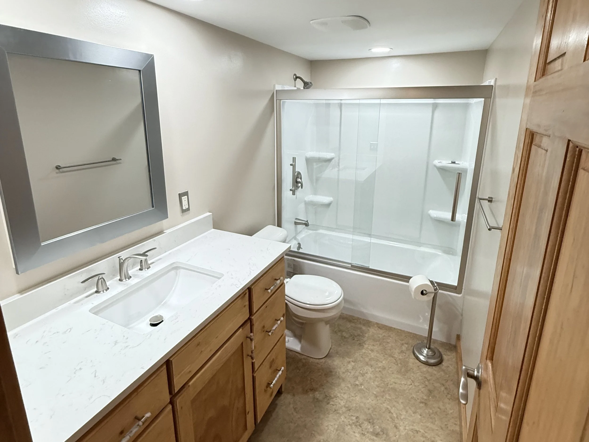 Bathroom with a wooden door, a vanity with a white countertop and sink, a mirror, a toilet, and a shower with glass doors and built-in shelves.
