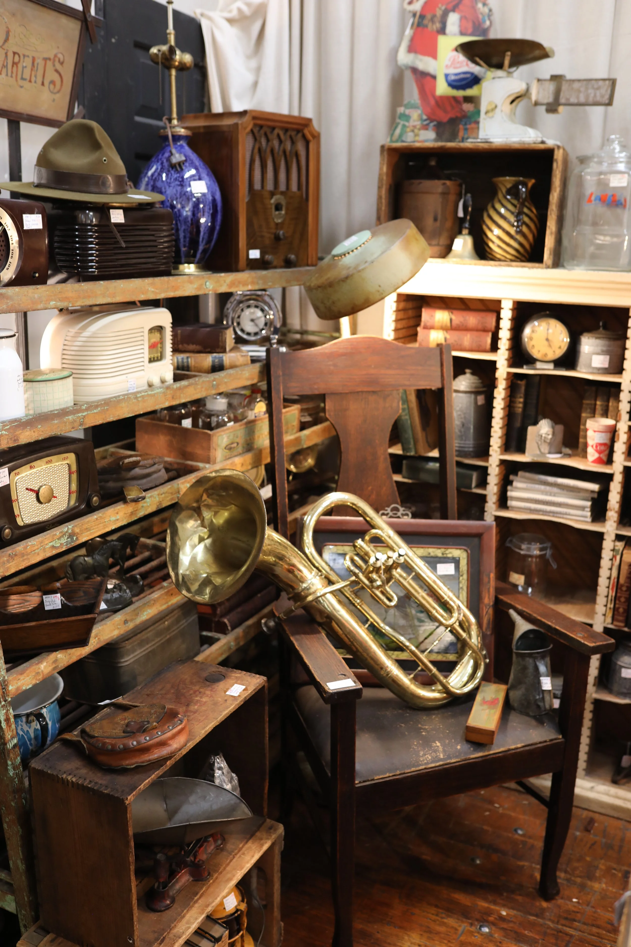 An antique shop filled with vintage items including a gold saxophone sitting on a wooden chair, old clocks, books, pottery, and various retro decorations.