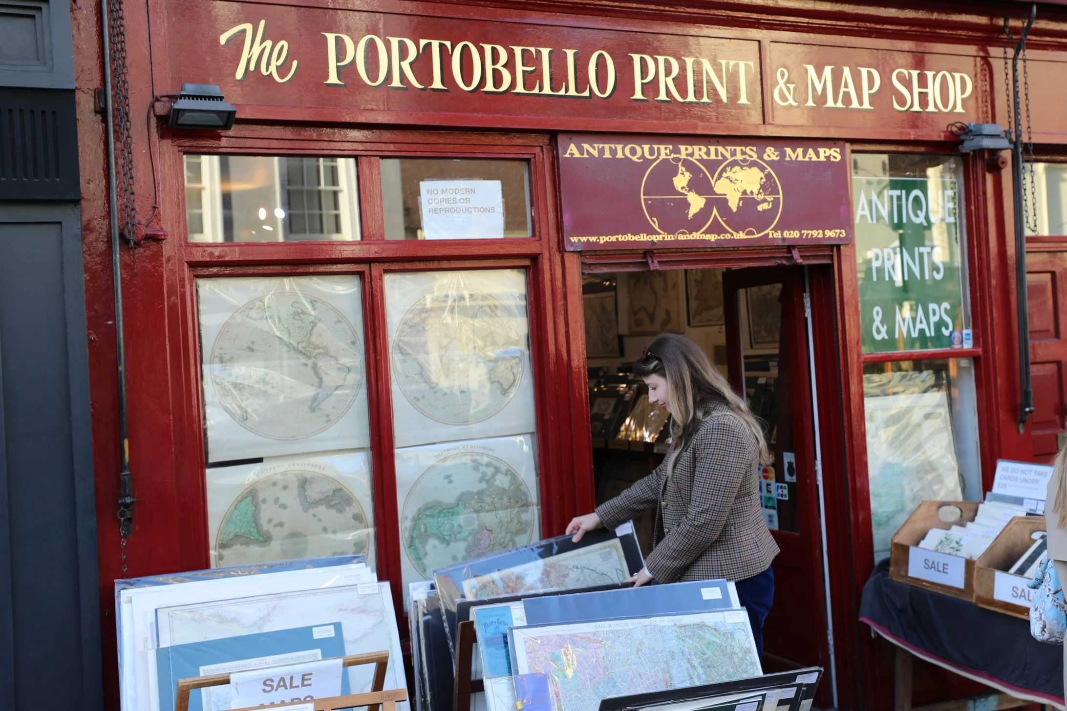 A woman browsing maps outside a shop called The Portobello Print & Map Shop, which sells antique prints and maps. The shop has large windows displaying various maps and a sign indicating a sale.