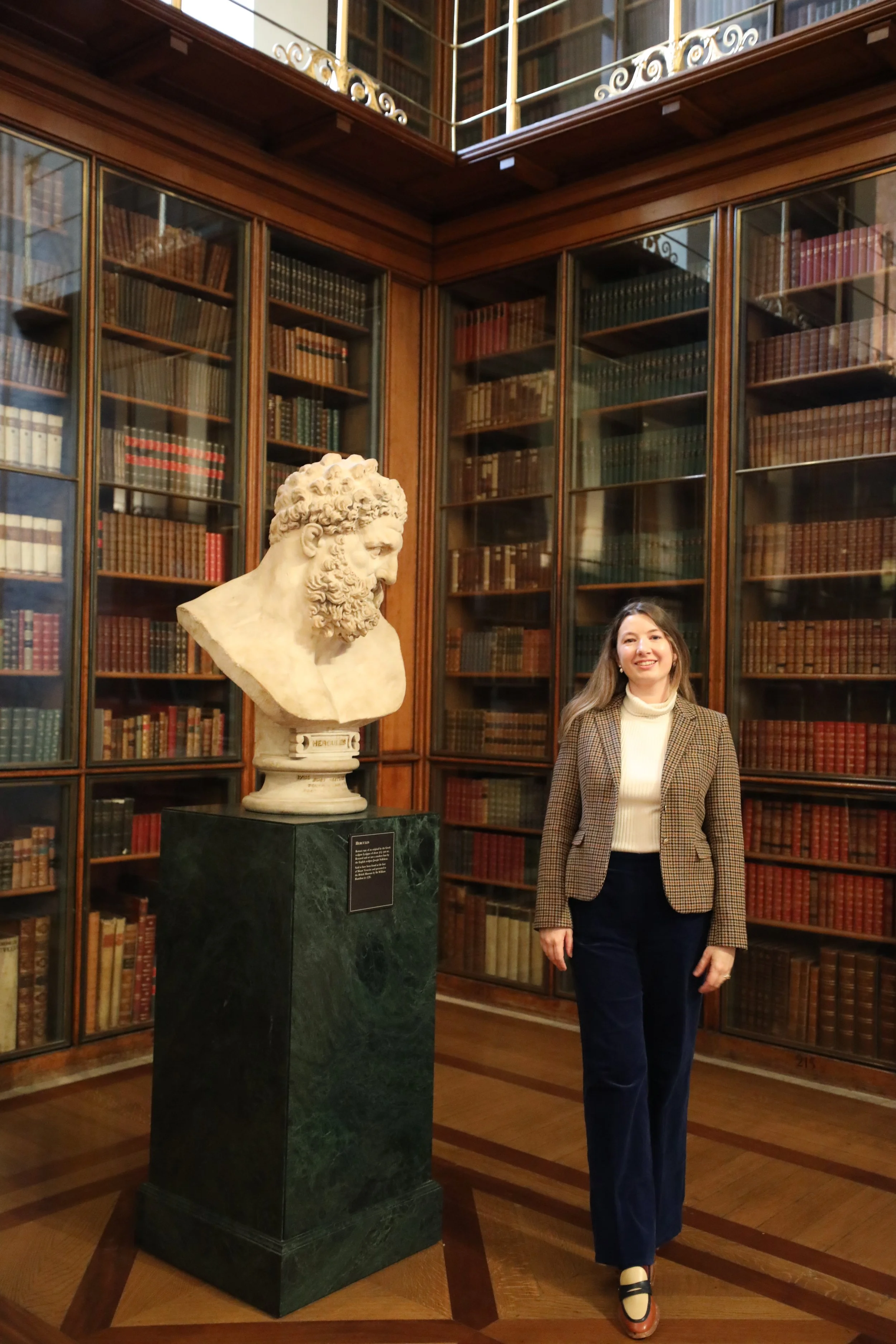 A woman standing inside a library with wooden bookshelves filled with old books, next to a marble bust sculpture of a bearded man on a pedestal.