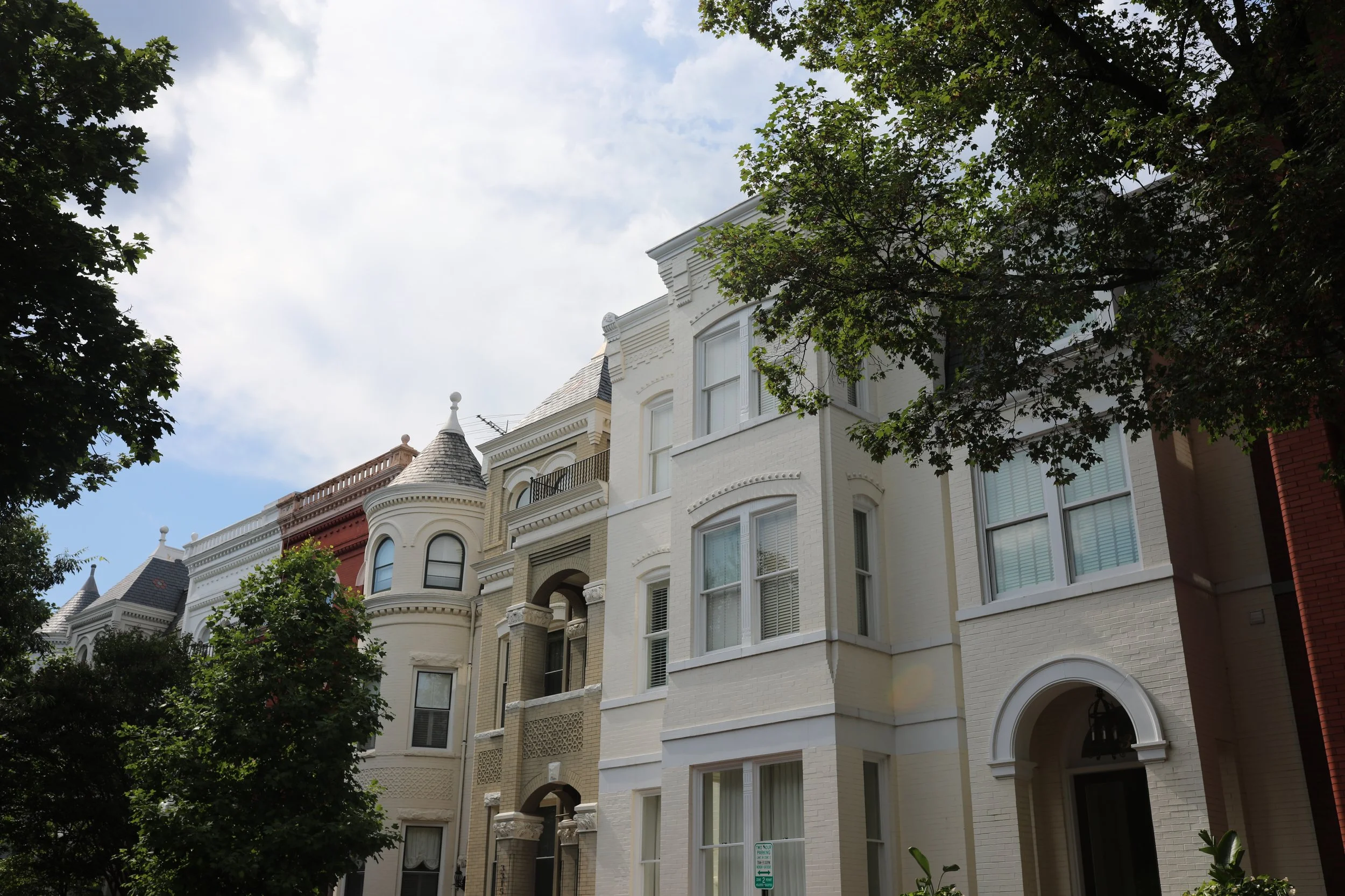 Historic white and beige row houses with decorative details, latticework, and turreted roofs, partly obscured by trees, under a partly cloudy sky.