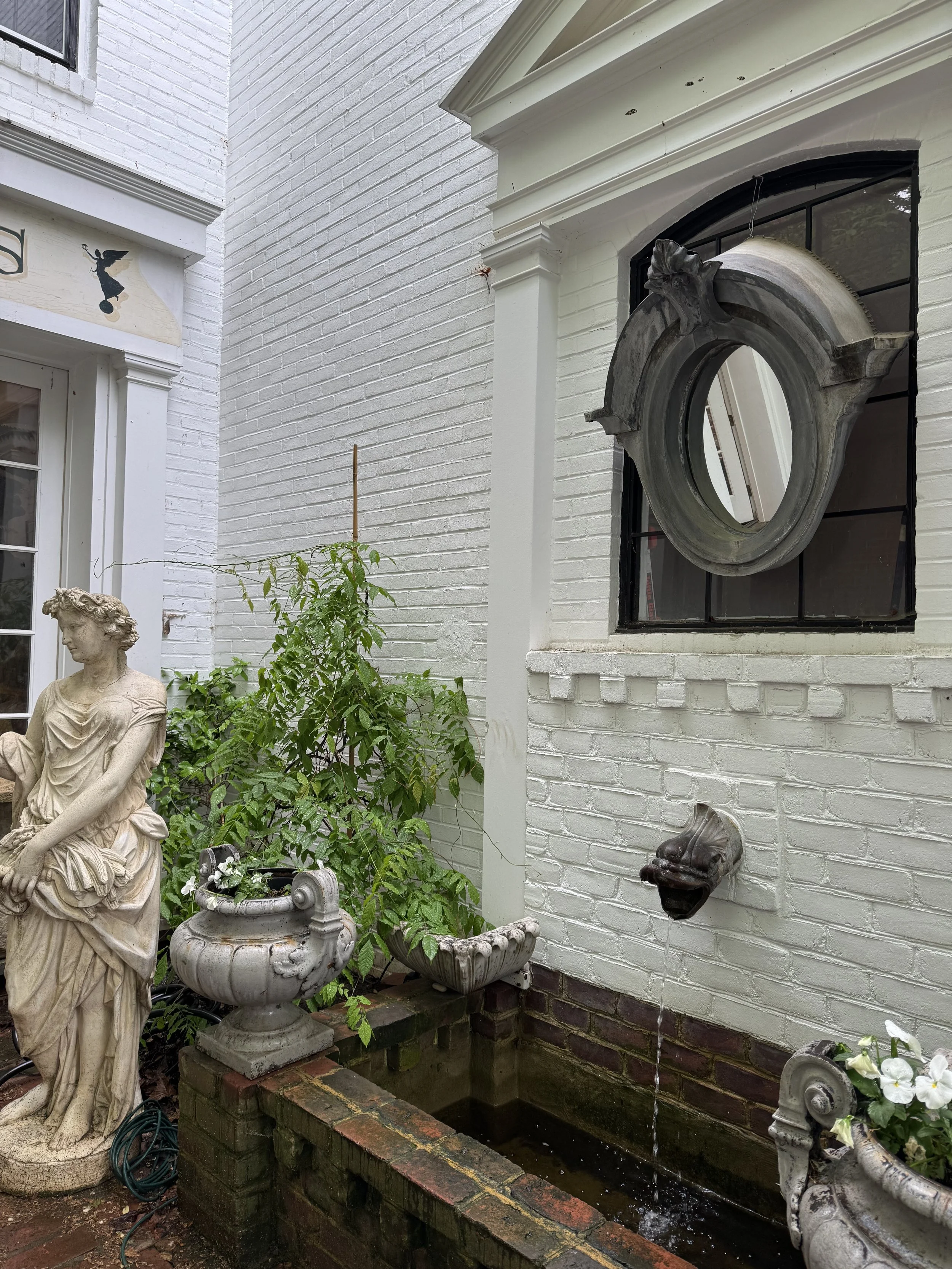 Exterior view of a white brick building with decorative elements, a garden statue of a woman, and a small fountain with water flowing from a spout.