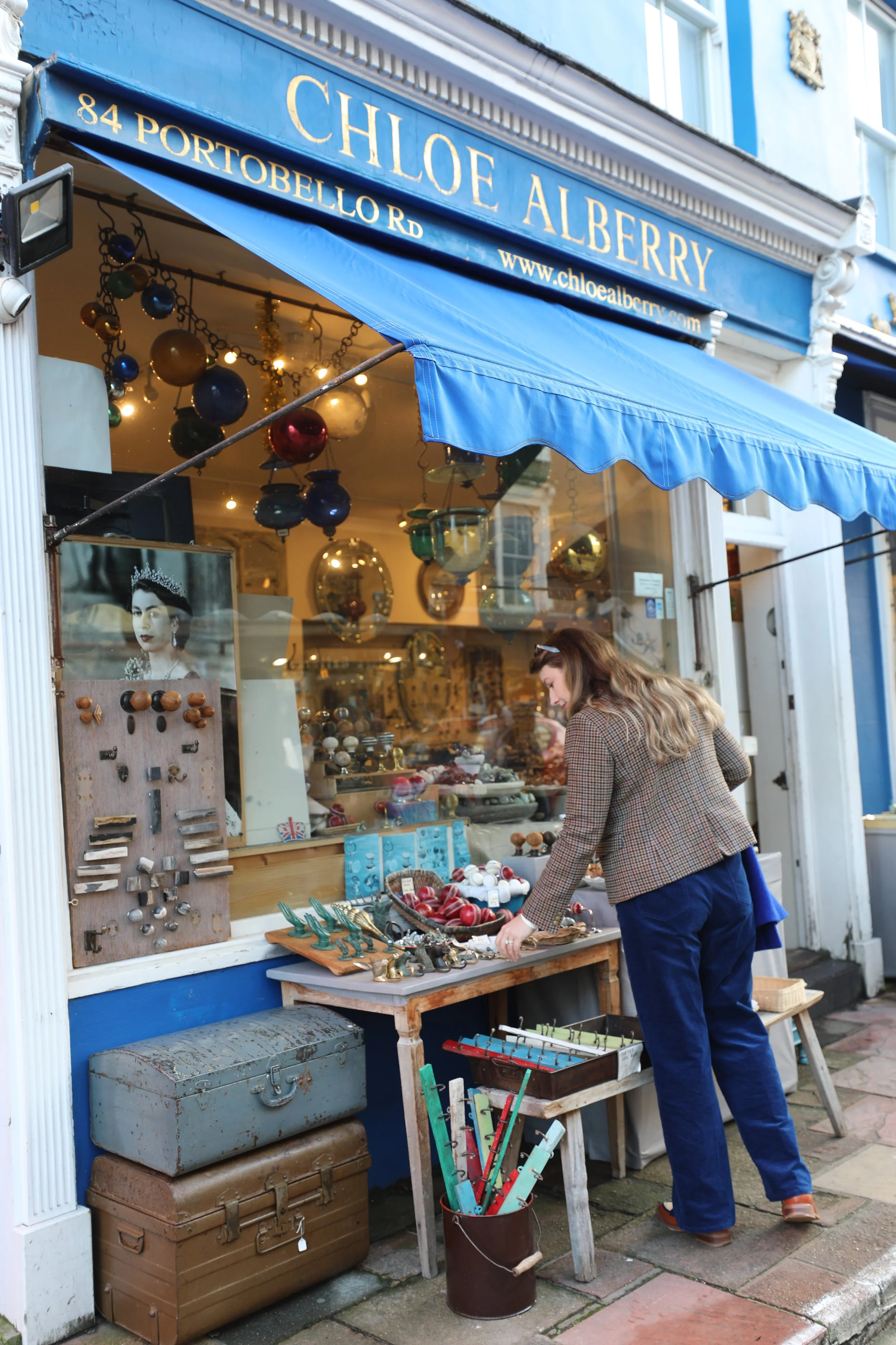 A woman browsing vintage collectibles outside Chloe Alberry store on Portobello Road, featuring glass ornaments, jewelry, and antique items in a shop window.